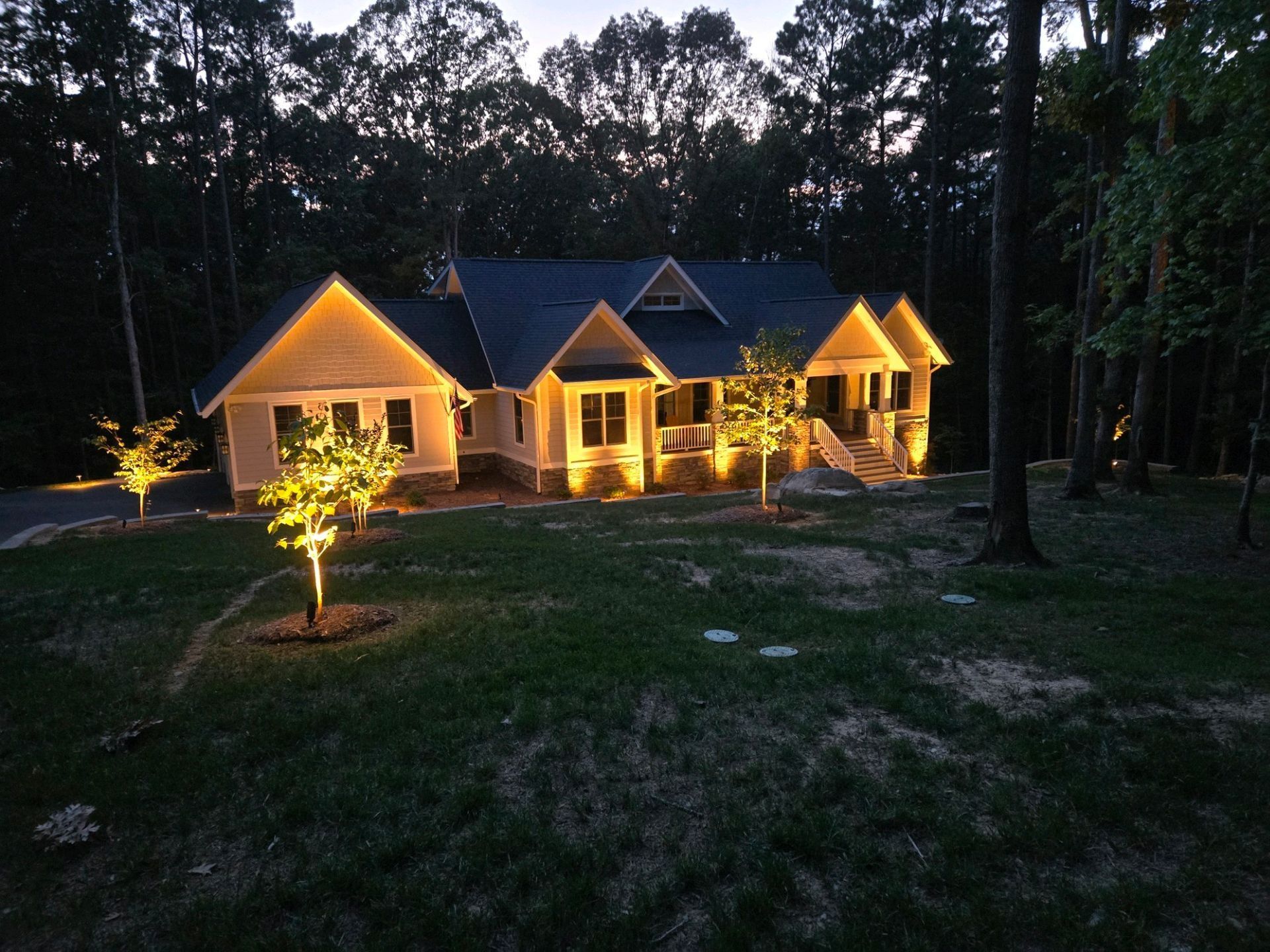 Warmly lit modern house at dusk, surrounded by tall trees and a grassy yard.