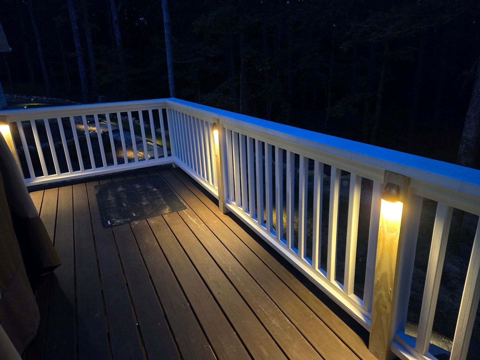 Dimly lit wooden deck with white railing and warm porch lights at night