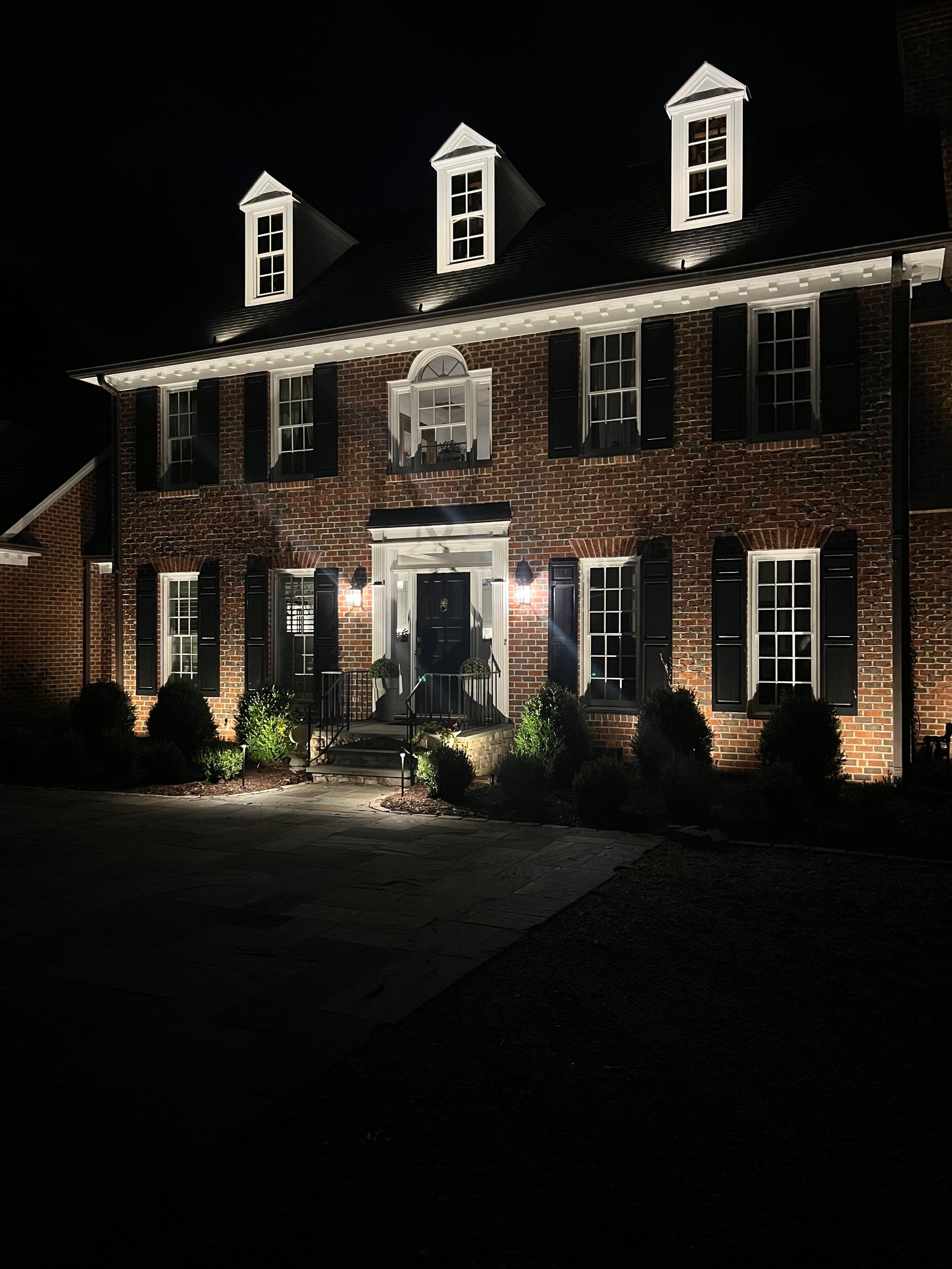 Brick house lit at night with white-trimmed windows and dormers, front entrance glowing warmly.
