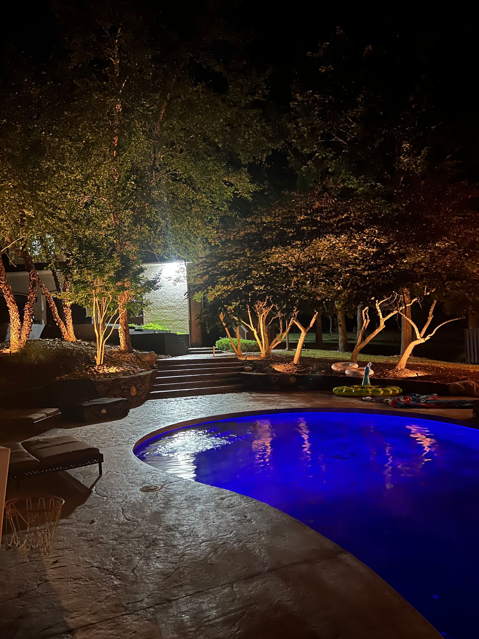 Nighttime pool lit blue beside a wet patio and trees, with a bright doorway in the background