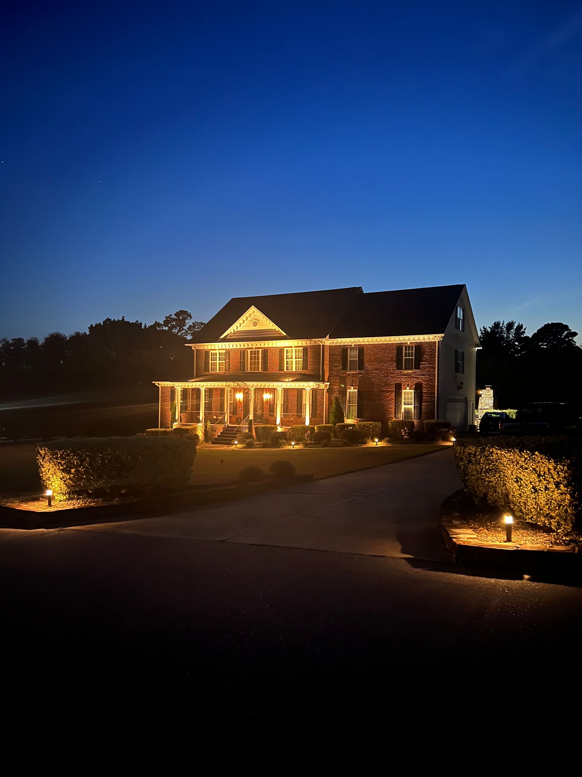 Lit house at dusk with warm porch lights and landscaped driveway under a deep blue sky