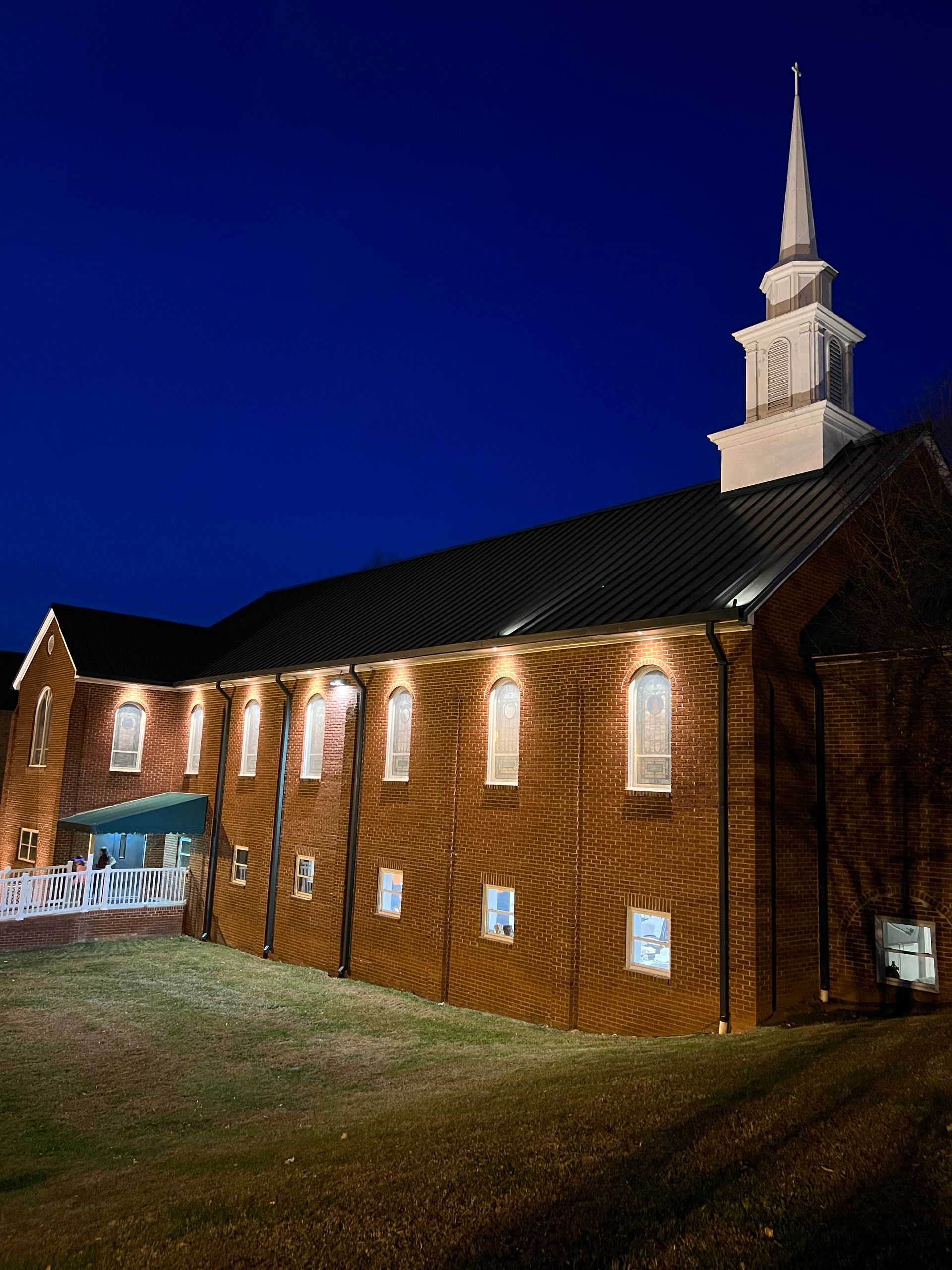 Brick church at night with lit windows and a tall white steeple against a dark blue sky