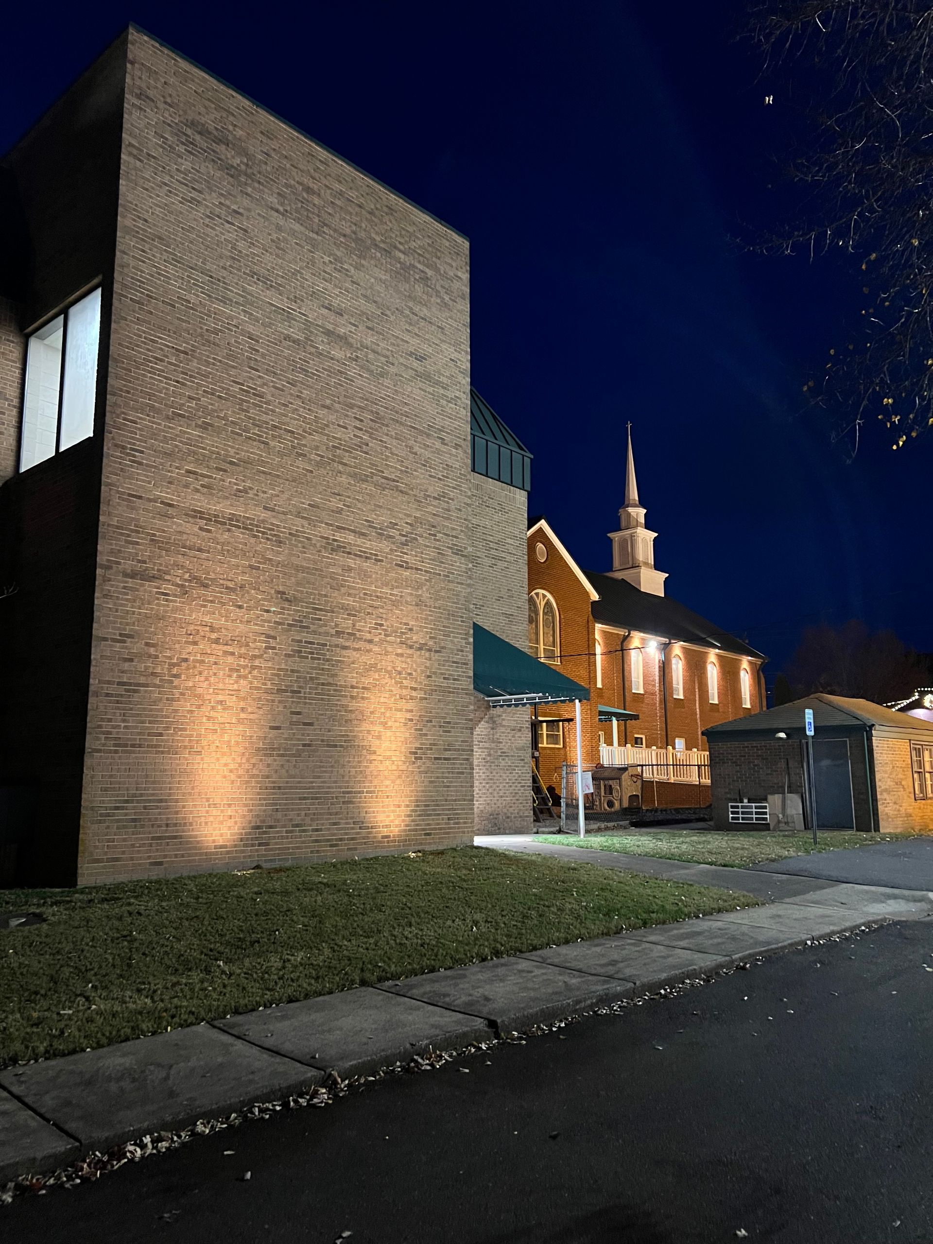 Nighttime view of a brick building and lit church tower beside a dark street and grassy verge