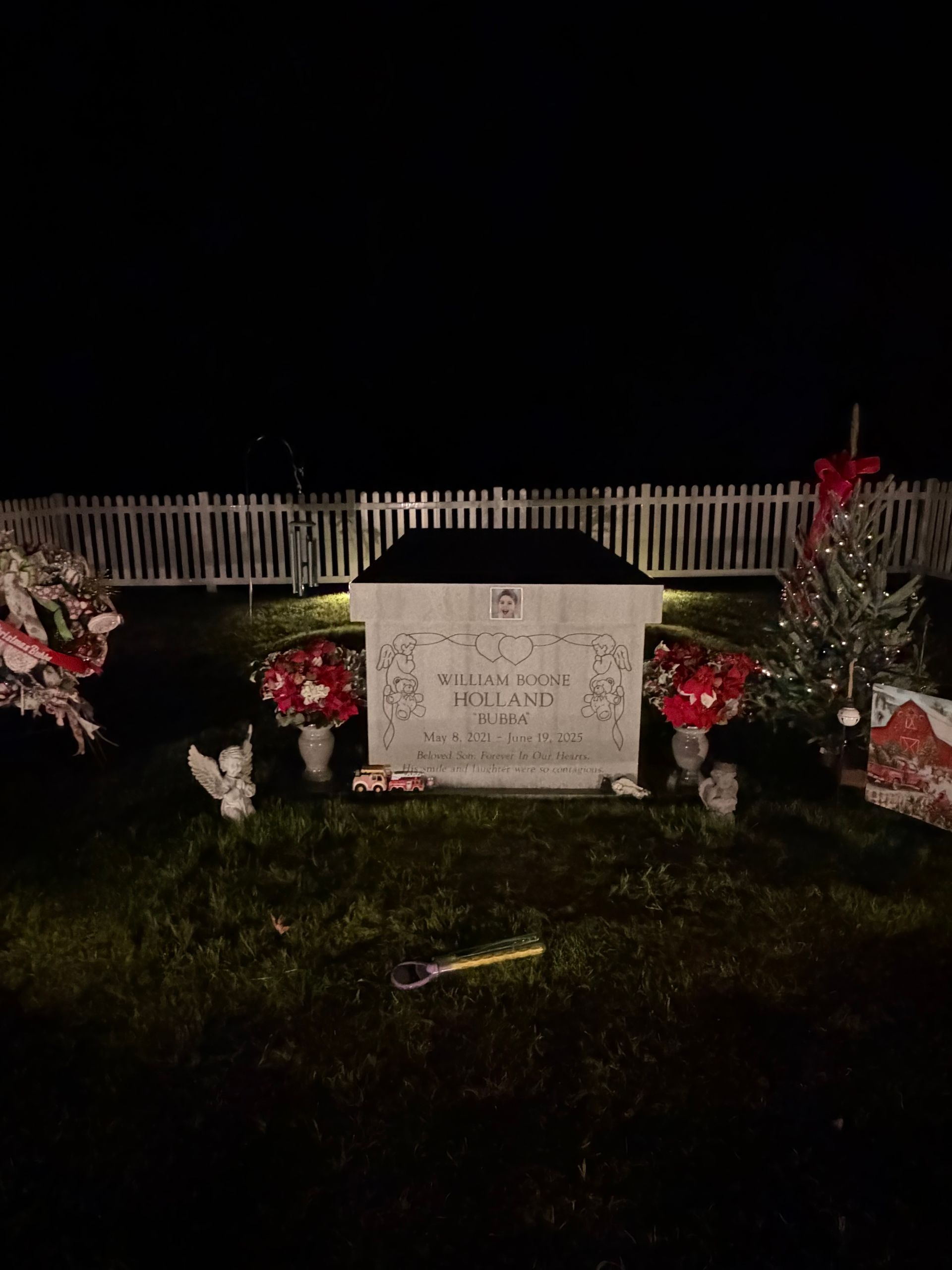 Nighttime grave with flowers and a stone marker in front of a white fence.