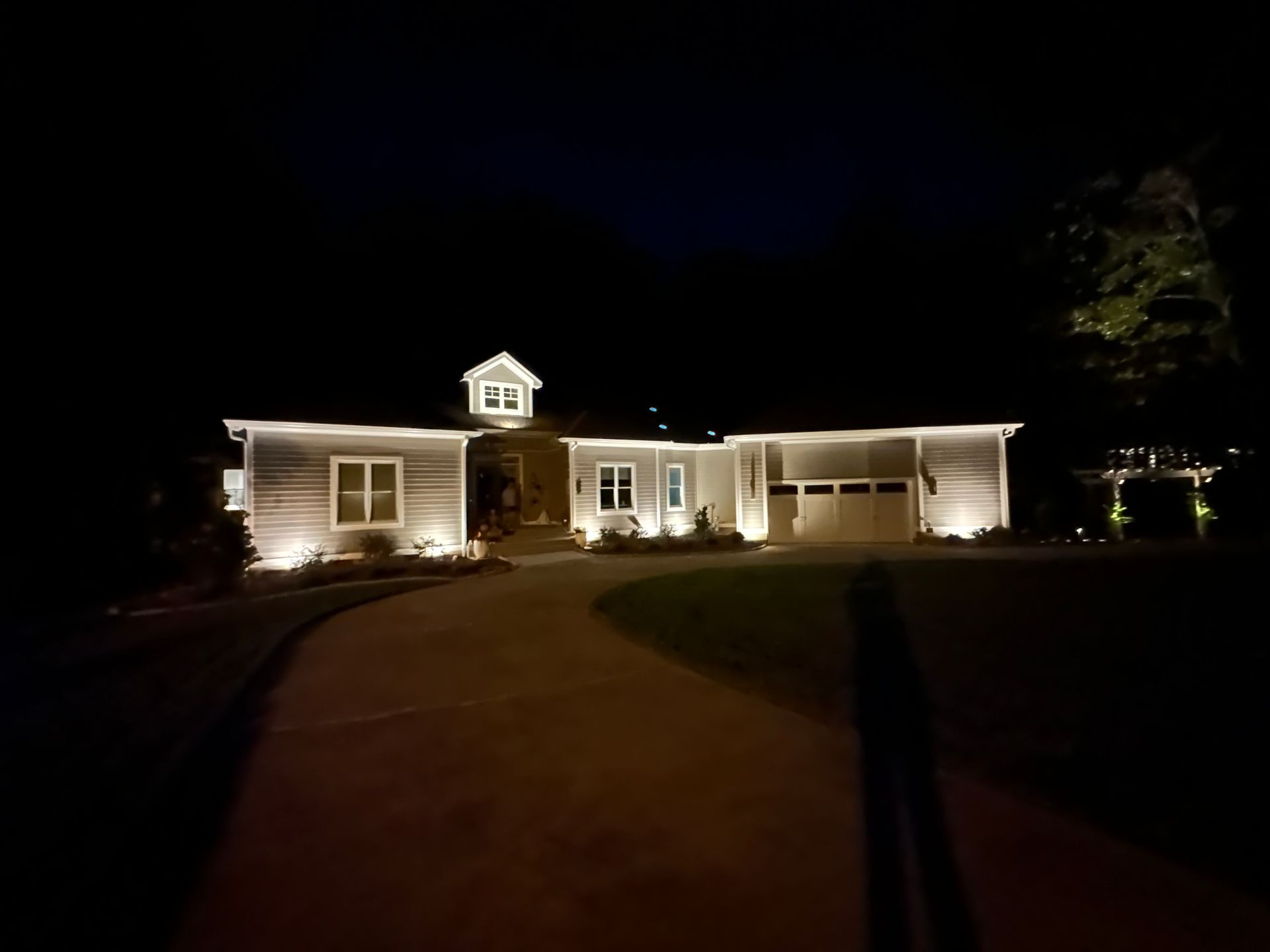 A house at night lit with warm white lights along the roof and windows, with a curved driveway in front.