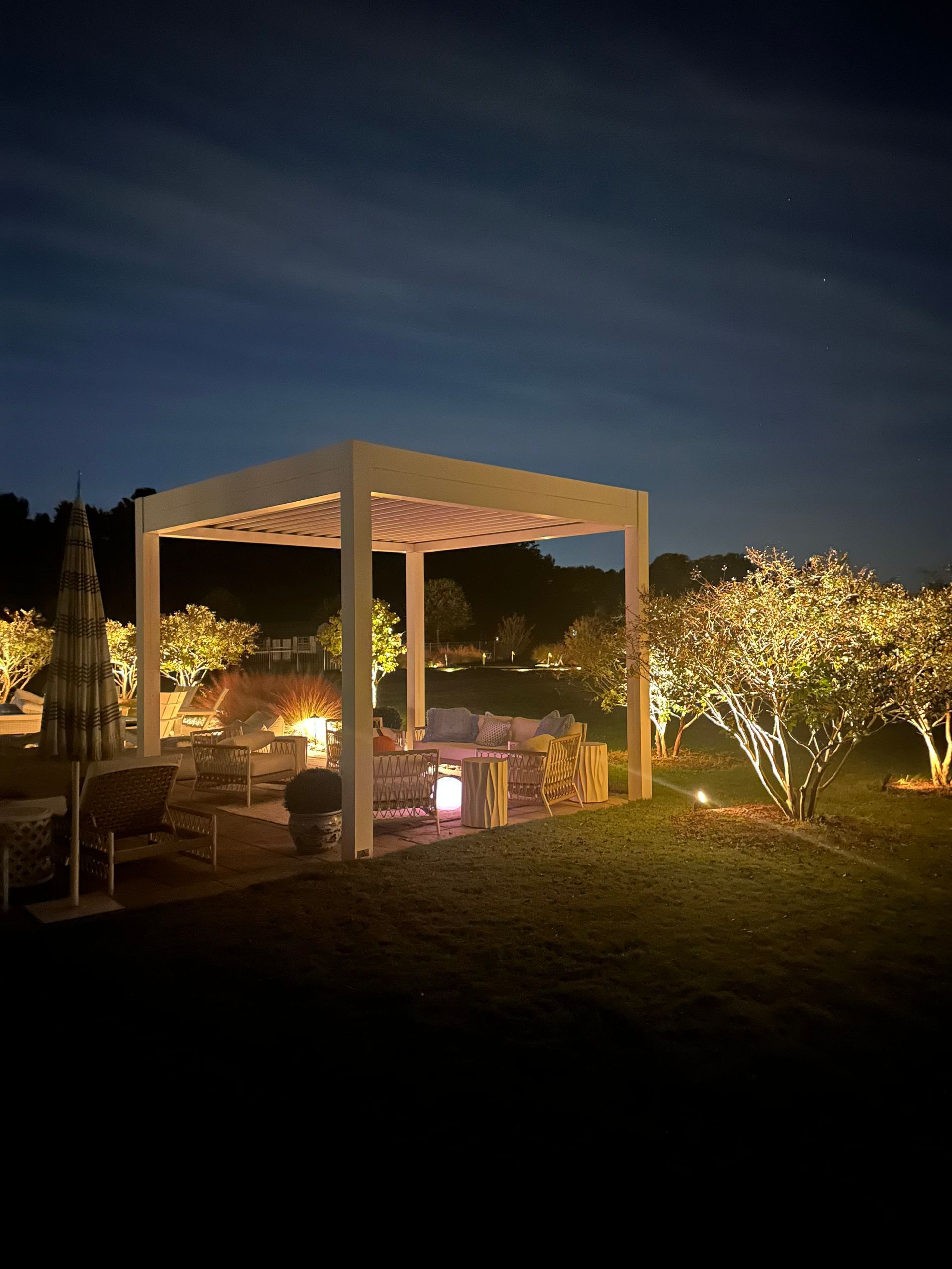 Nighttime garden patio with a white pergola, warm lights, and illuminated trees under a dark sky