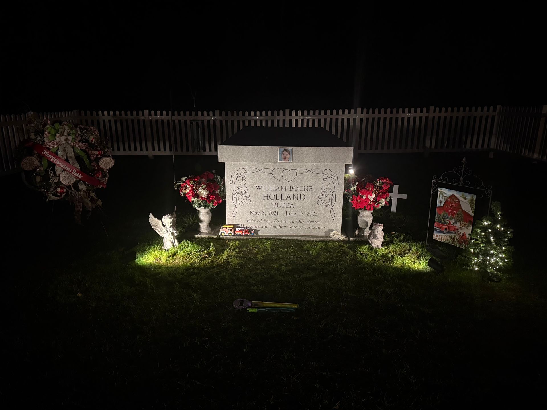 A nighttime memorial with flowers and lights around a decorated gravestone by a fence.