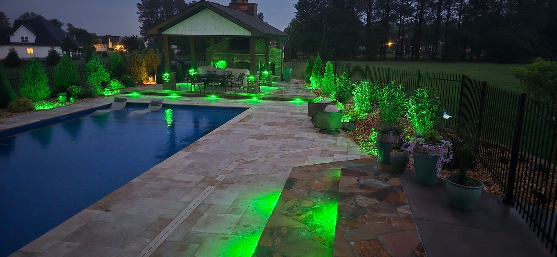 Backyard pool at dusk with green landscape lighting and a patio leading to a lit pavilion