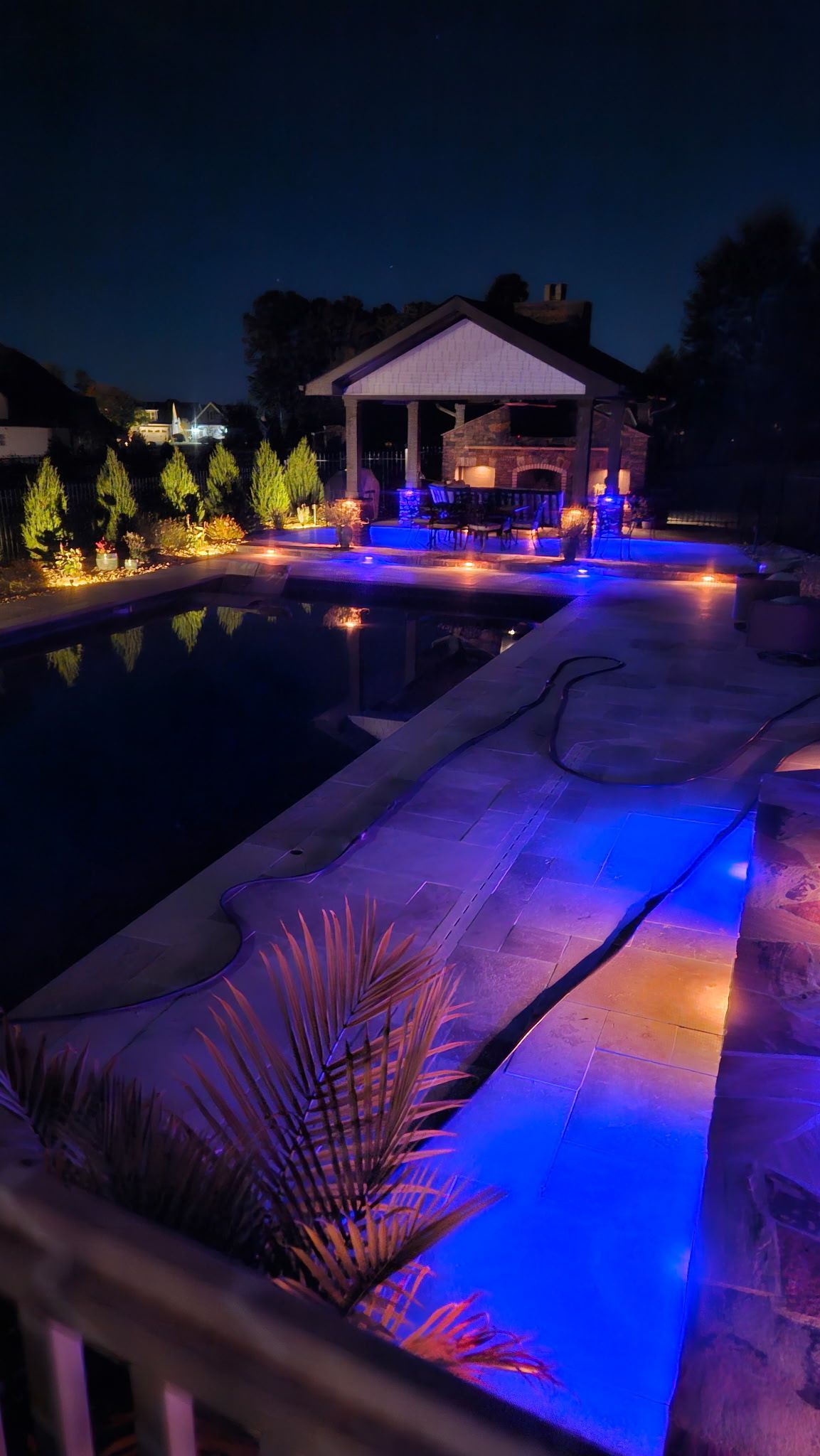 Nighttime poolside patio with blue-purple lights and a small pavilion beside the water