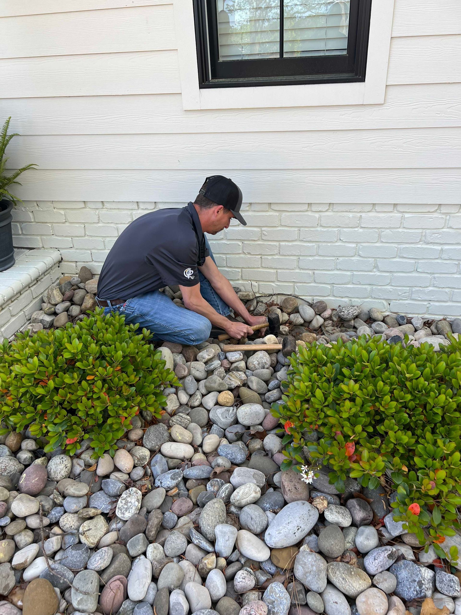 Person kneeling in a rock garden, working near a white brick house wall and black window.
