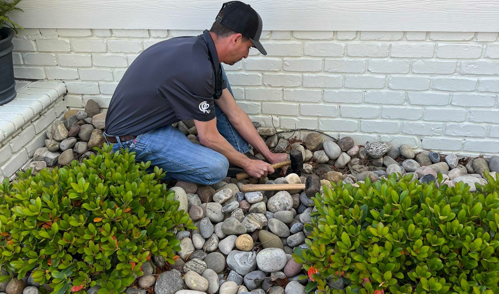 Man kneels in a rock garden, using a hammer to place stones near a white brick wall.