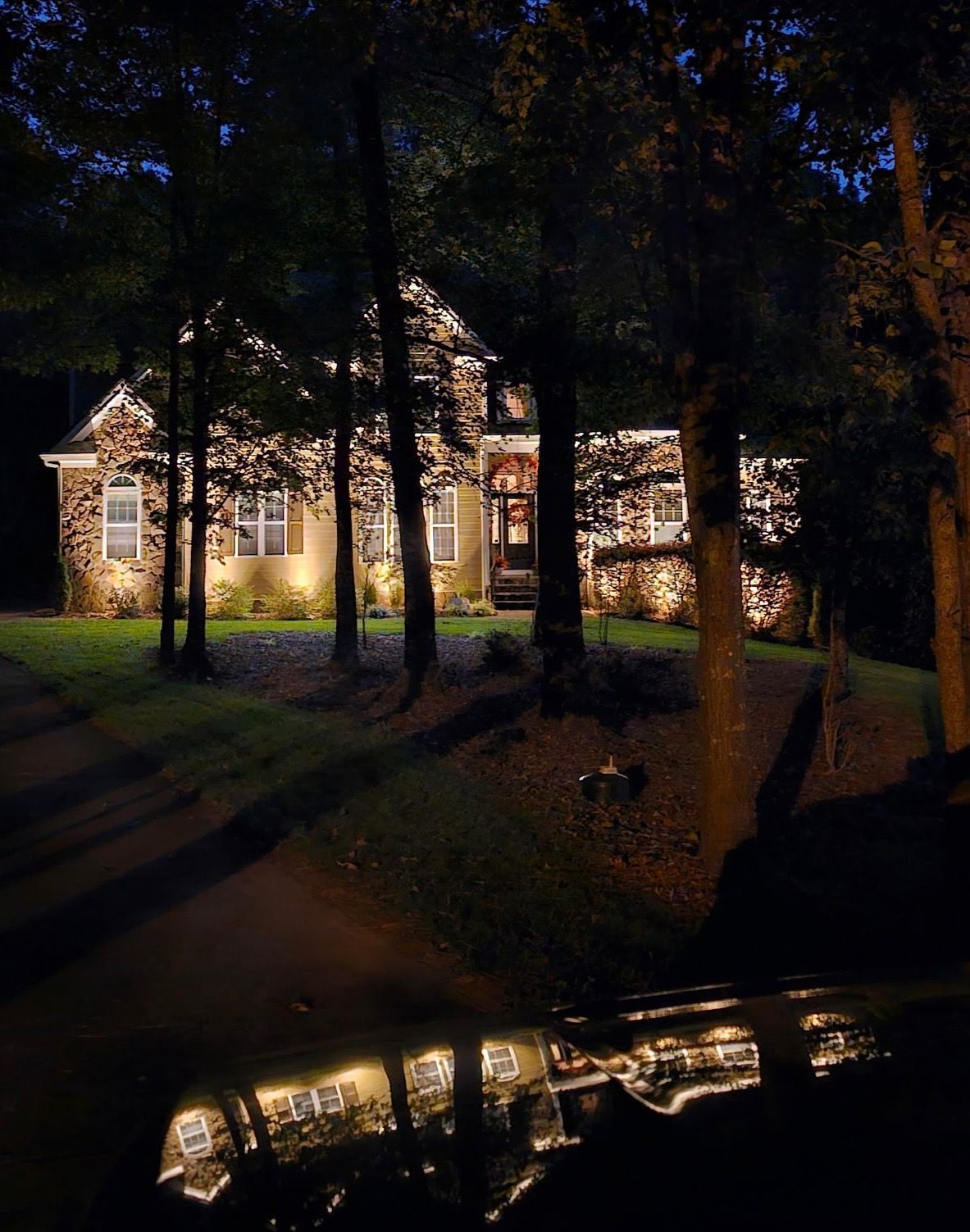 Illuminated house at night behind trees, with lights reflecting on a car hood in the foreground