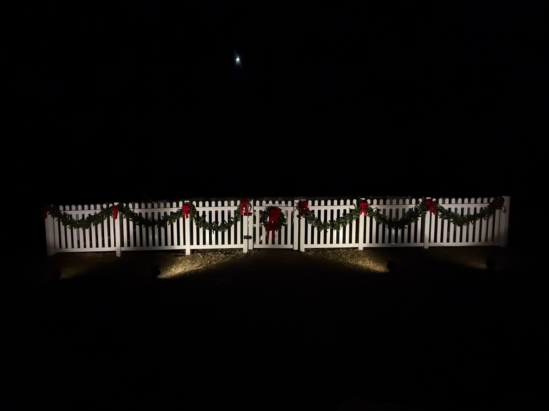 White fence decorated with red bows and greenery lit at night against a dark sky