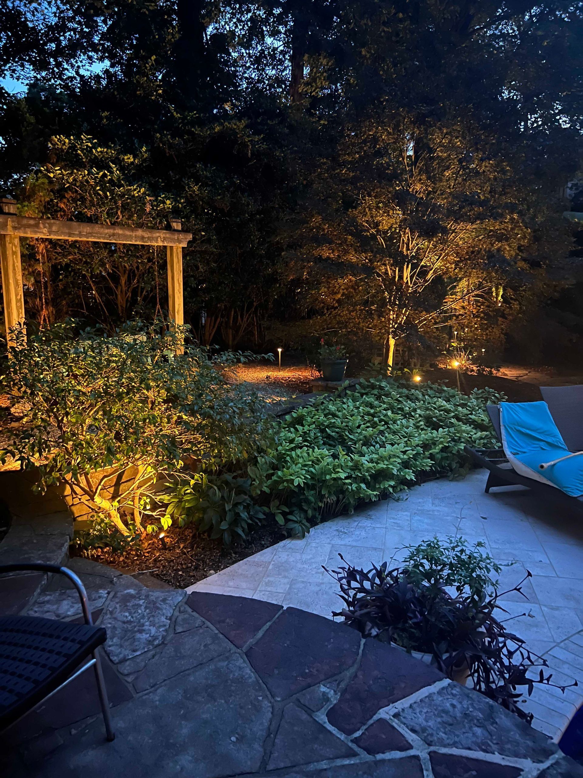 Backyard patio at night with warm string lights, stone walkway, lush greenery, and a blue lounge chair