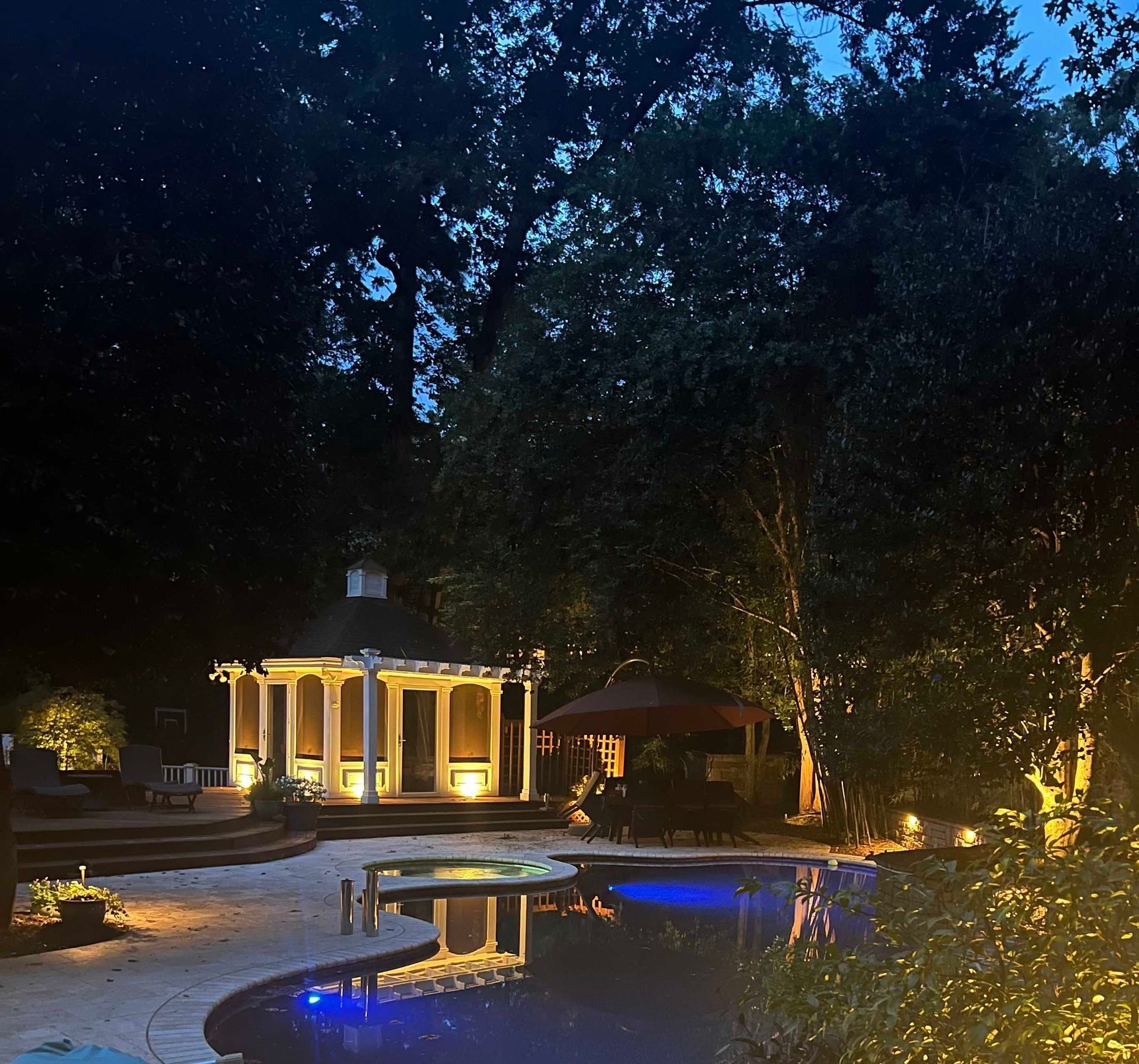 Nighttime courtyard with a lit gazebo, glowing pool, and tall trees silhouetted against the dark sky
