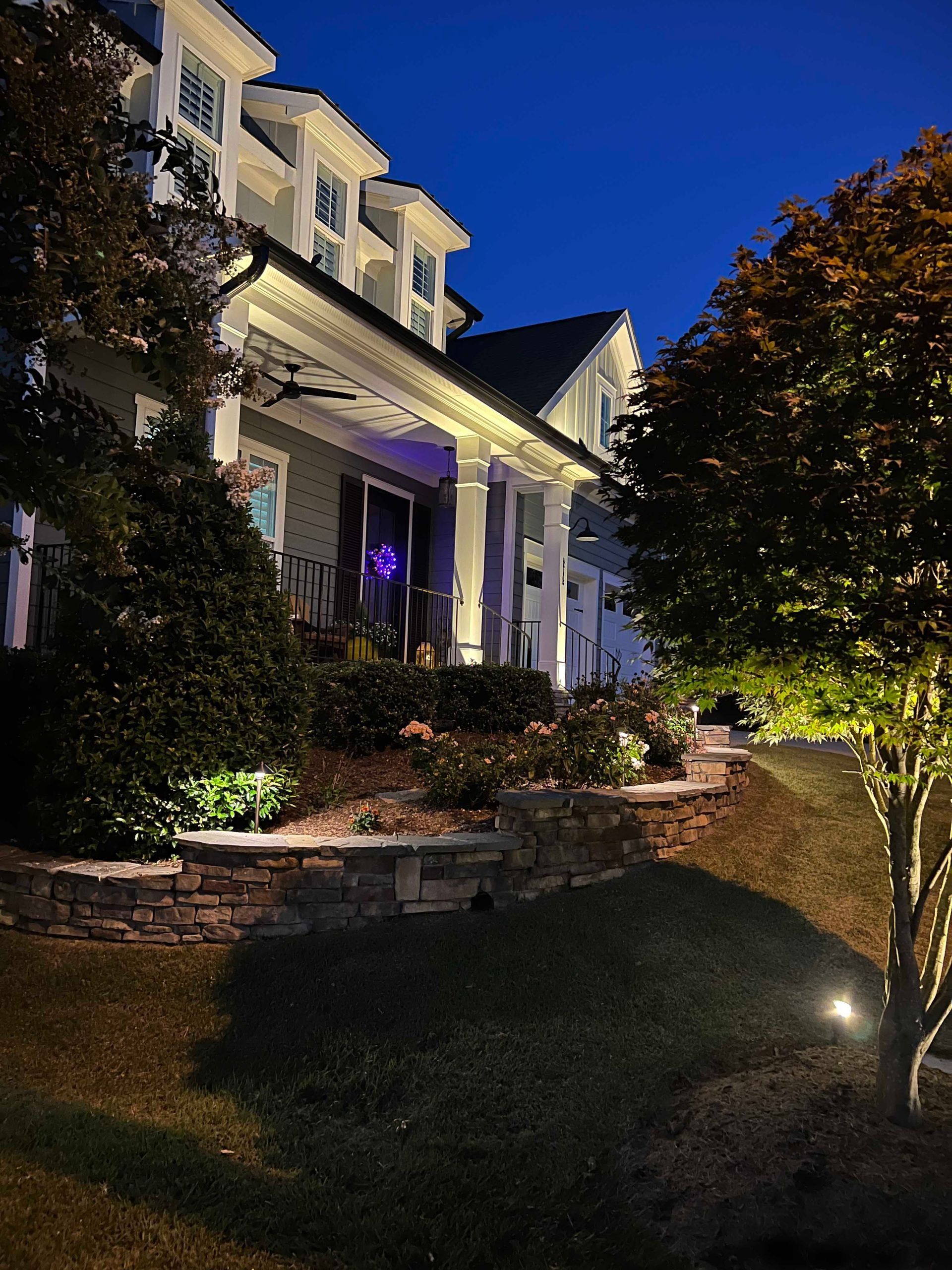 Lit house exterior at night with white porch columns, glowing path lights, and landscaped shrubs