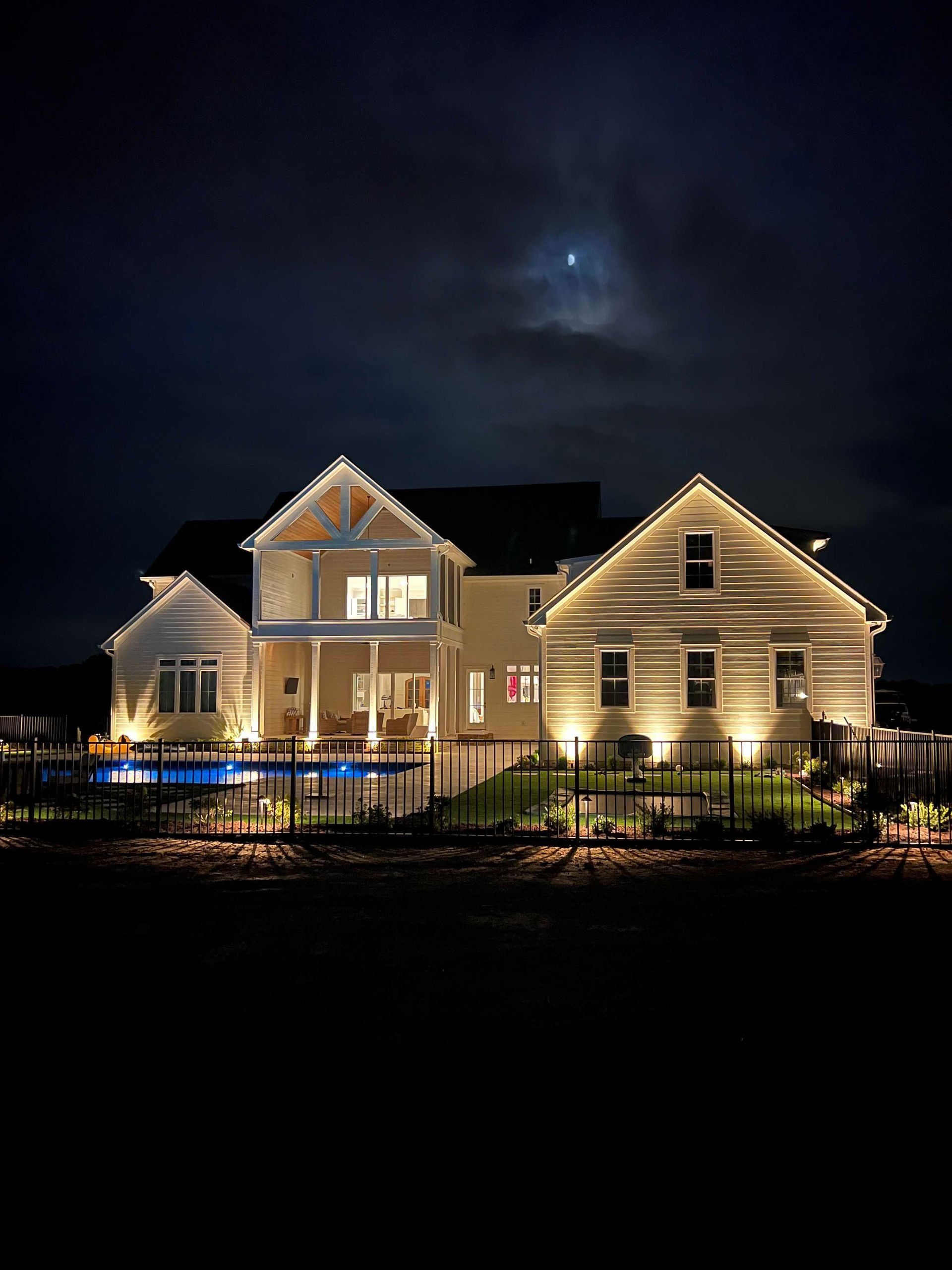 Illuminated two-story house at night with a lit pool and moonlit sky
