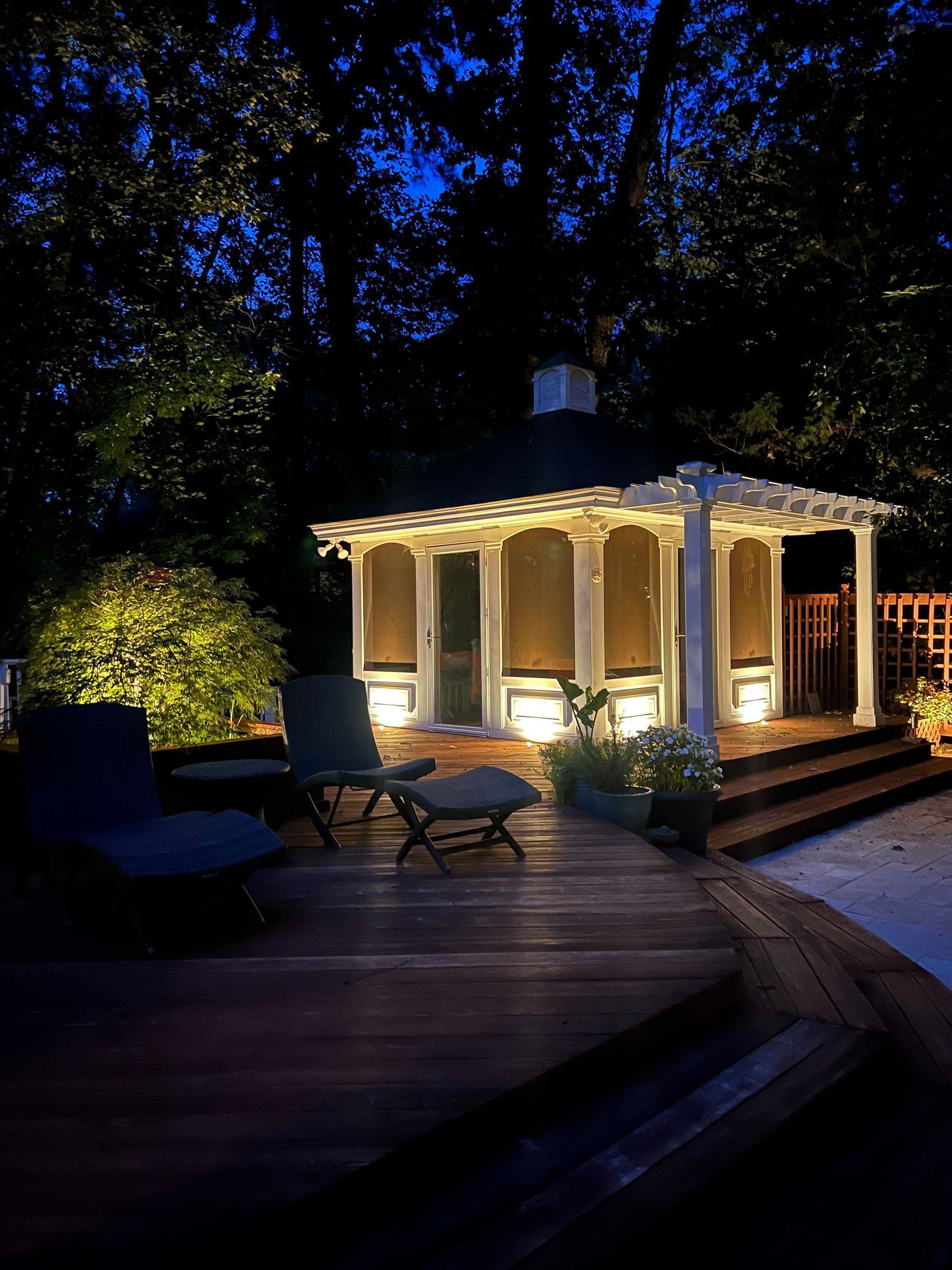 Lit backyard gazebo with string lights on a dark deck at night, surrounded by trees and patio chairs