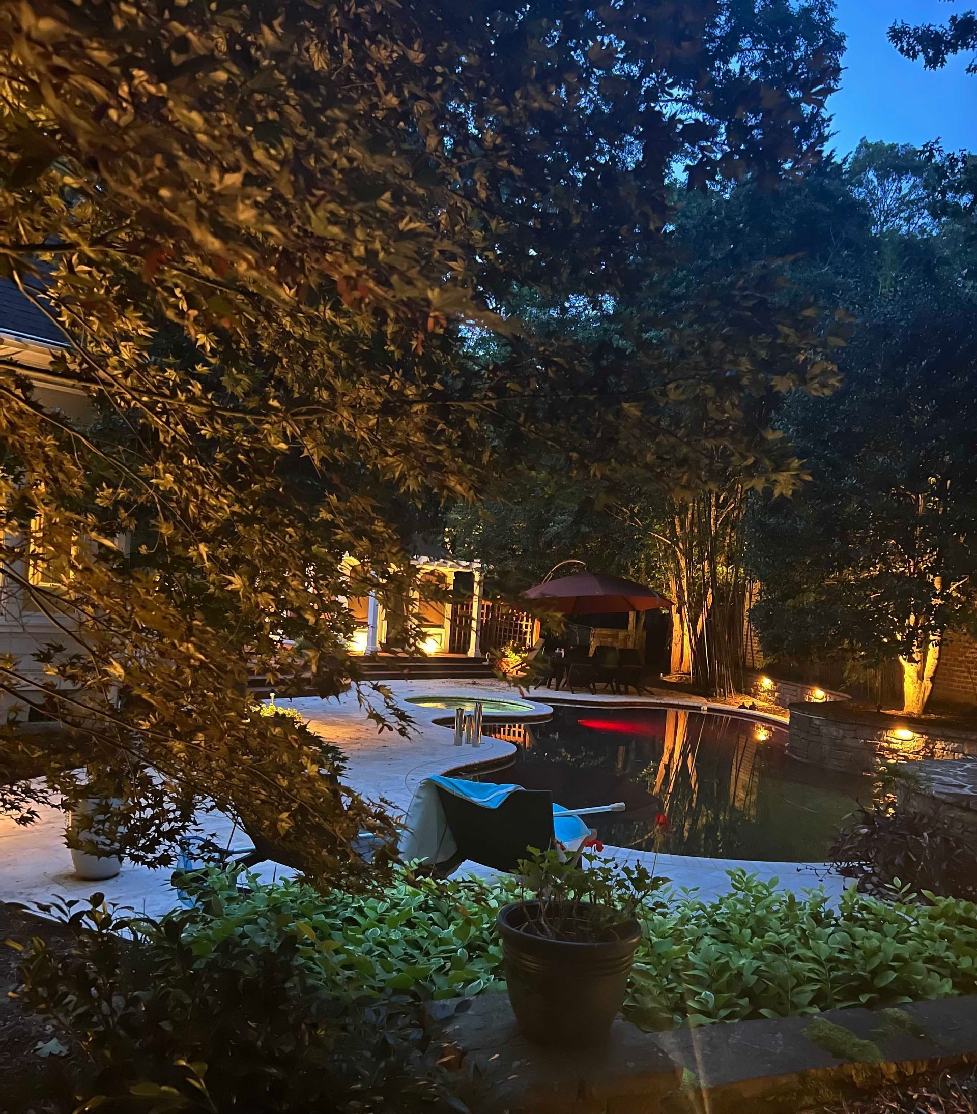 Nighttime backyard with lit trees, a pool, and a small table by the water.
