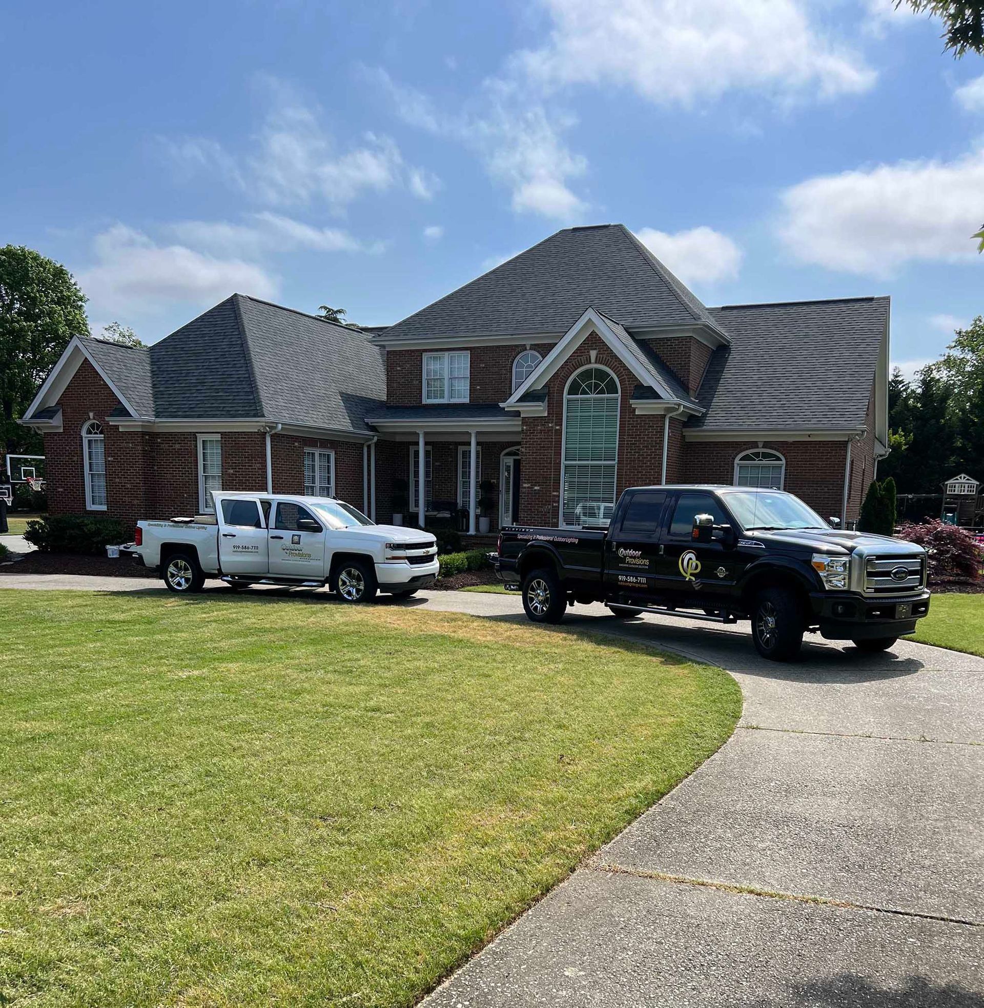 Brick house with a curved driveway, green lawn, and two pickup trucks parked out front under a blue sky