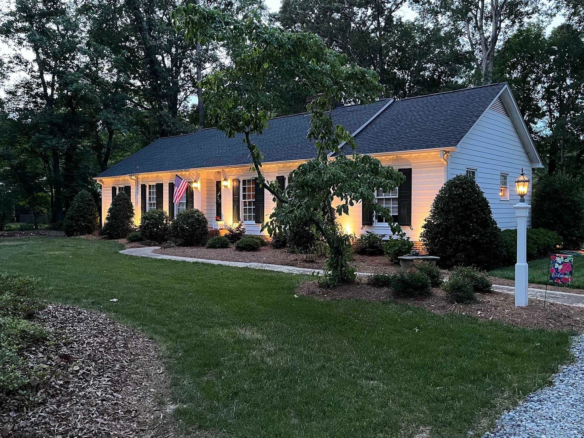 Single-story white house with lit porch lights, dark roof, and landscaped yard at dusk
