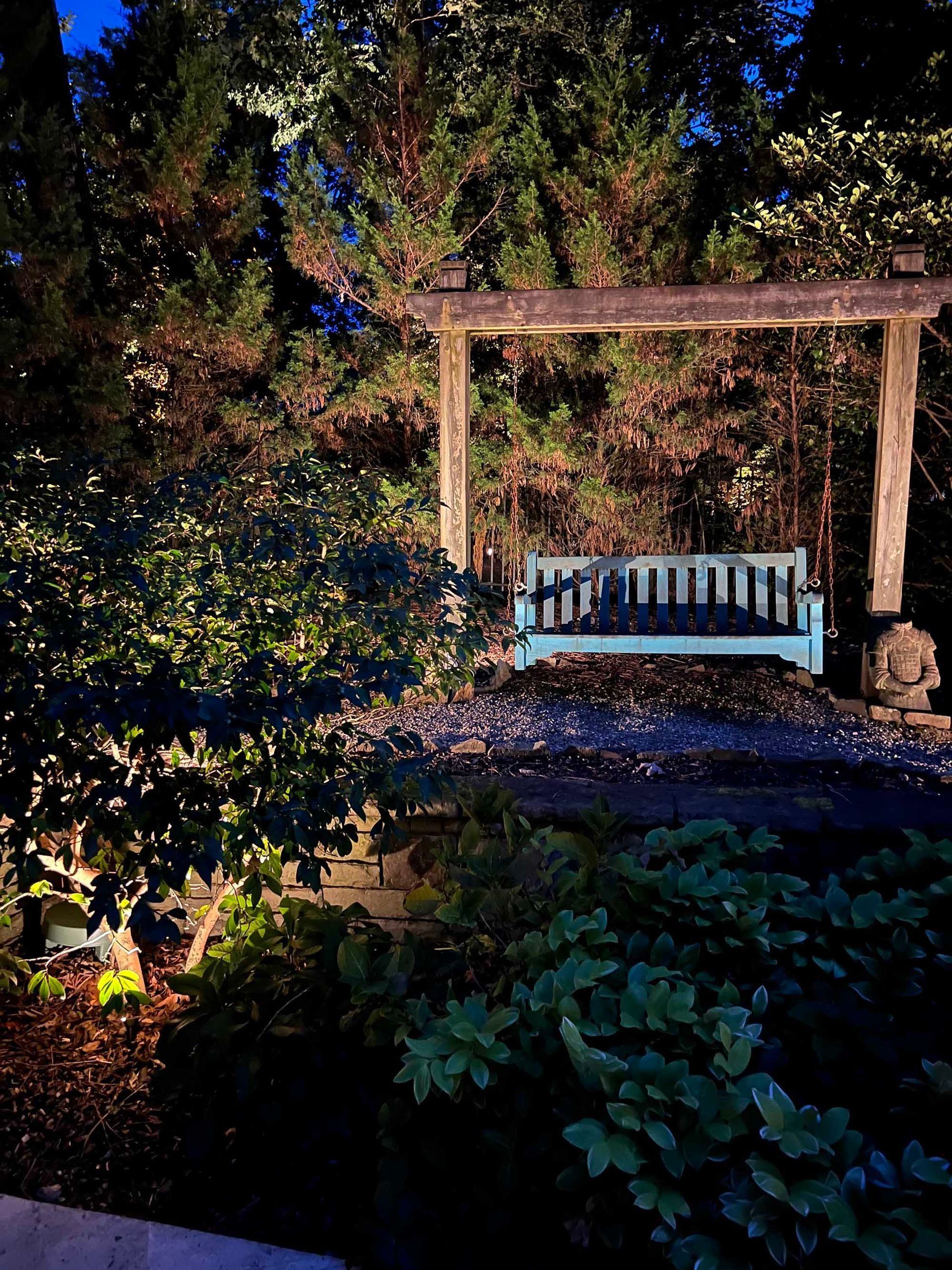 White bench under a wooden pergola in a sunlit garden, surrounded by trees and shrubs.