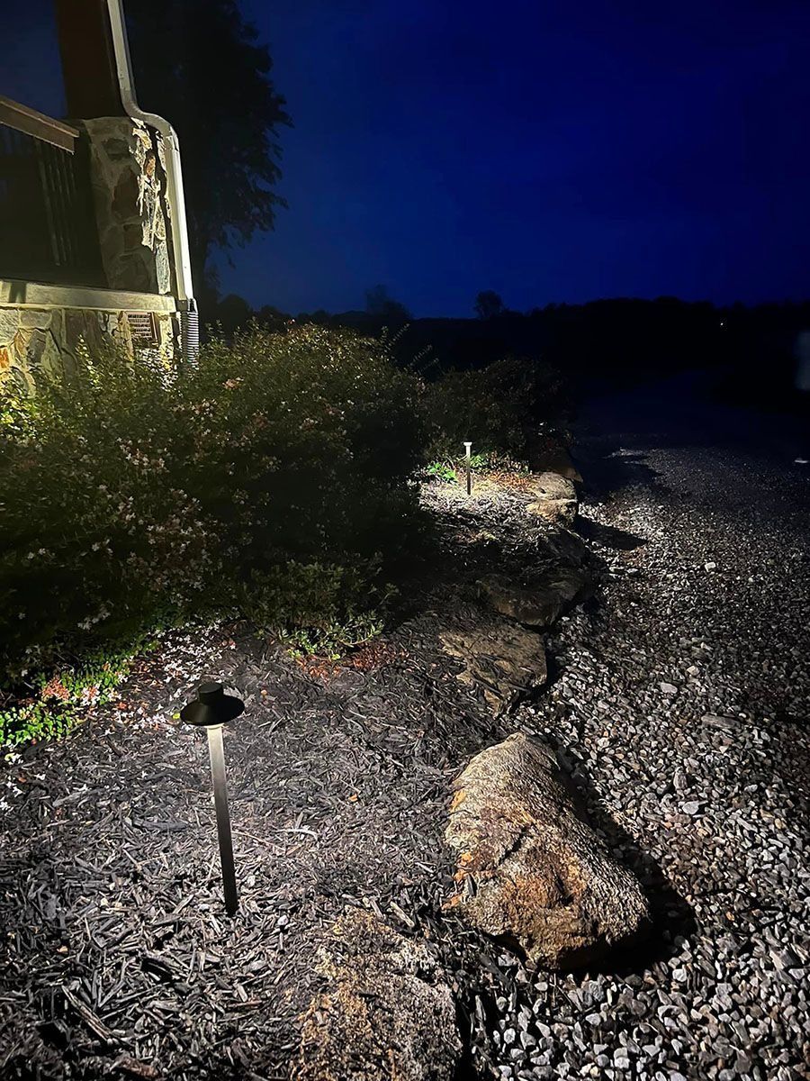 Nighttime gravel path beside a lit house and shrubs, with a large rock in the foreground.