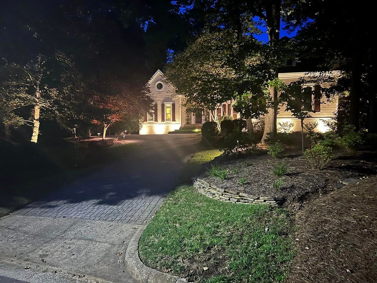 Nighttime view of a house with lit windows, a gravel driveway, and trees in the foreground.