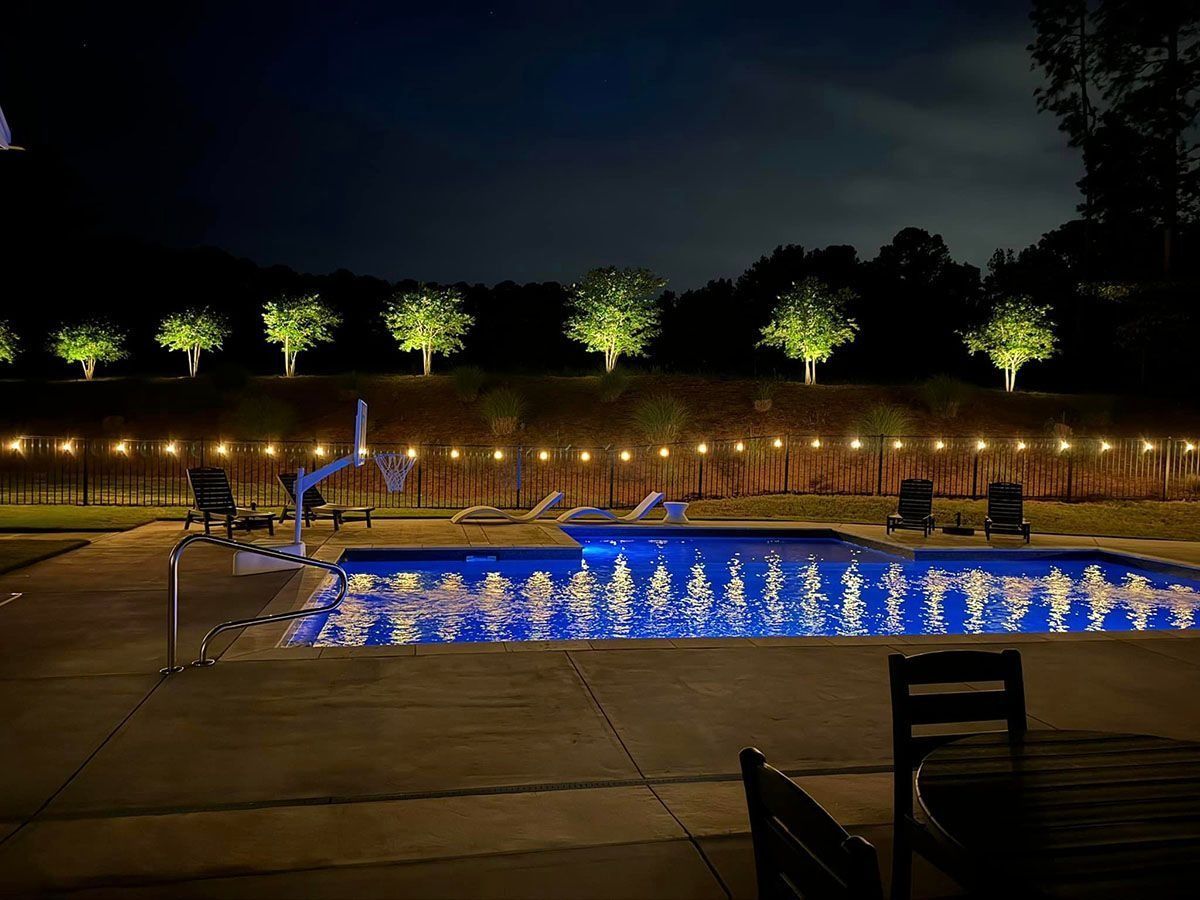 Illuminated outdoor pool at night with blue lights, lounge chairs, and trees along the fence