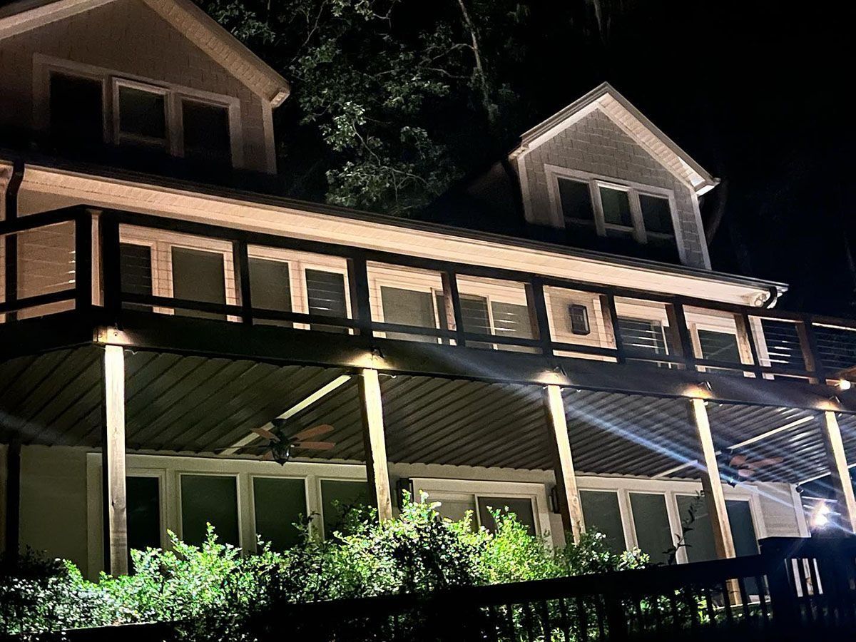 Lit house with wraparound porch and dormers at night, viewed from below amidst dark trees and shrubs
