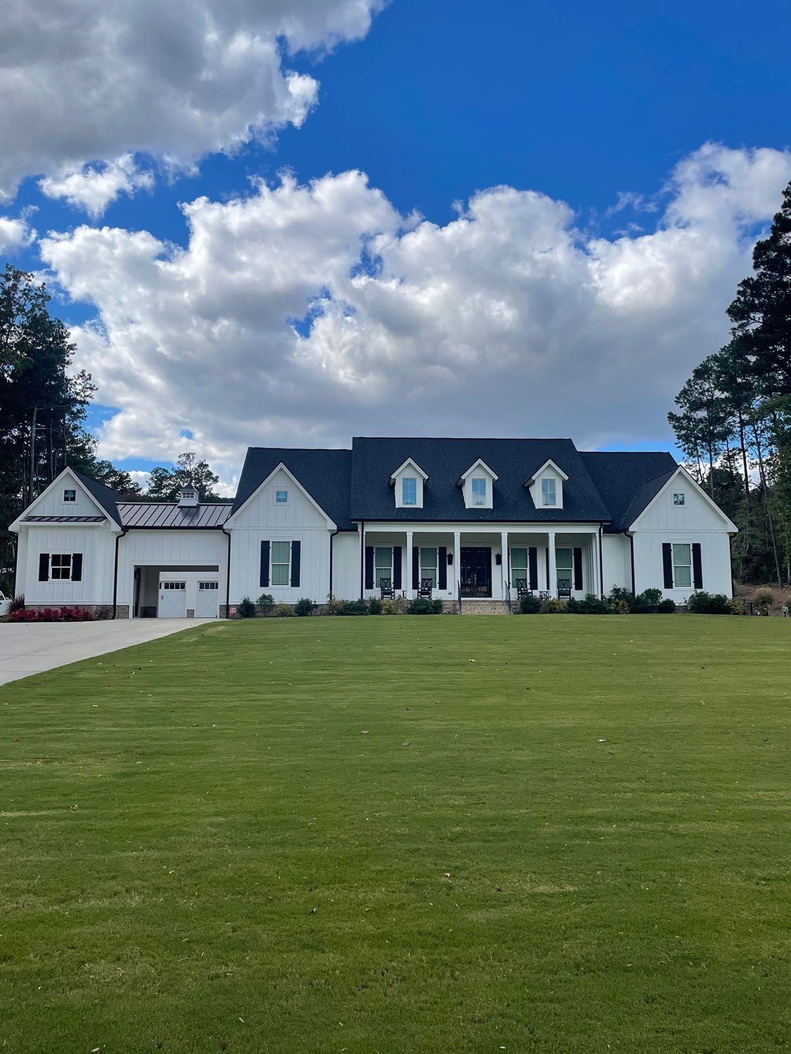Large white farmhouse with dark roof and porch under a blue sky with clouds, viewed across a green lawn