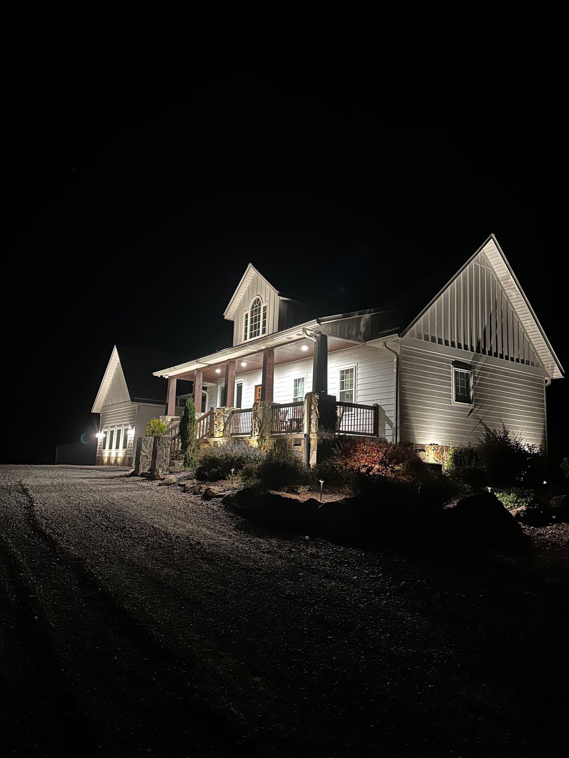 Illuminated white house at night with porch lights and dark gravel driveway