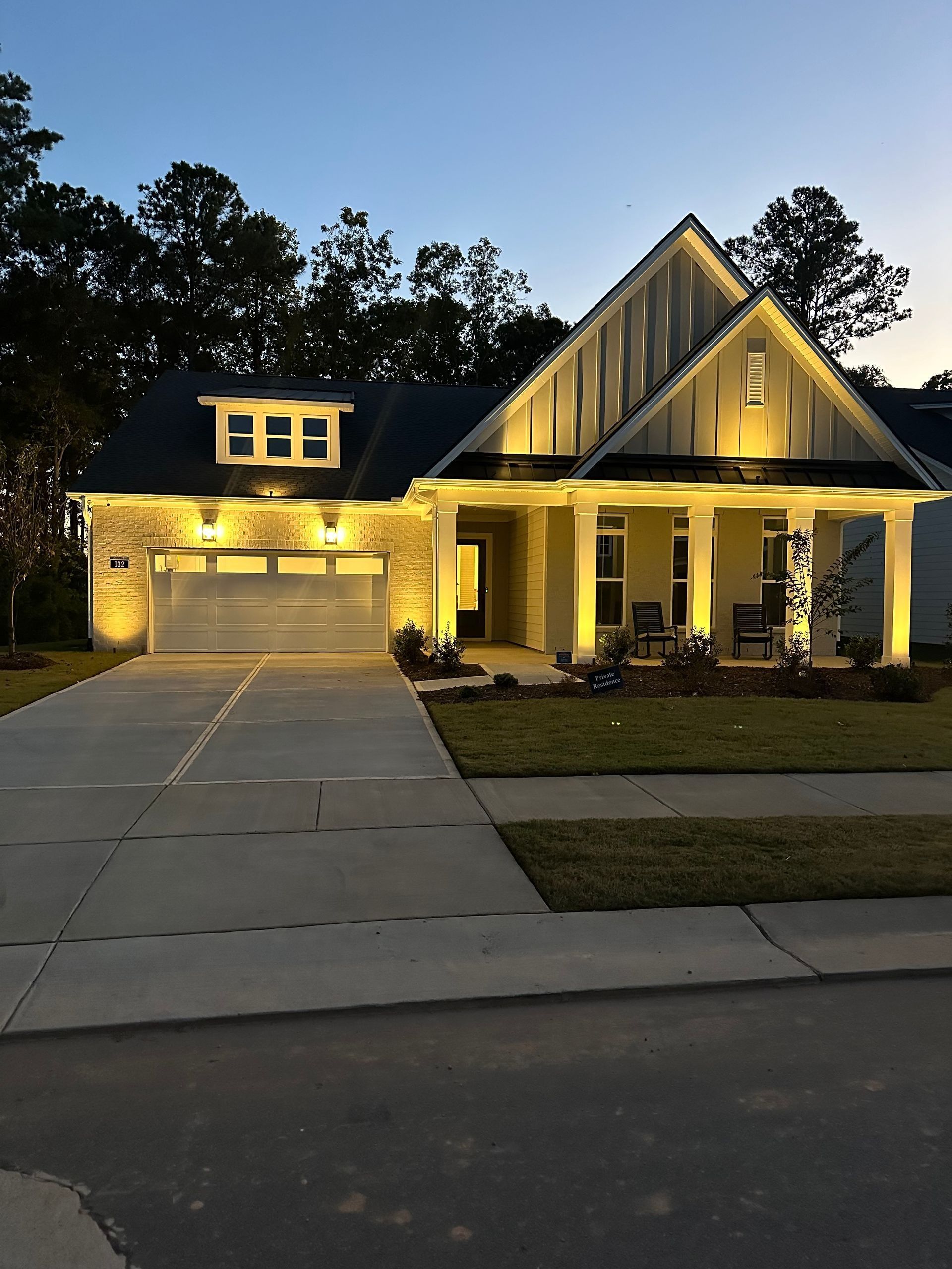 Suburban house at dusk with warm porch lights, lit garage, and driveway in front of tall trees
