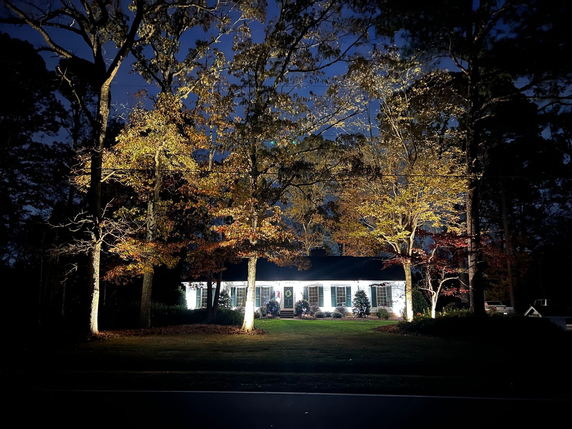 Night view of a lit white house behind autumn trees and a dark lawn