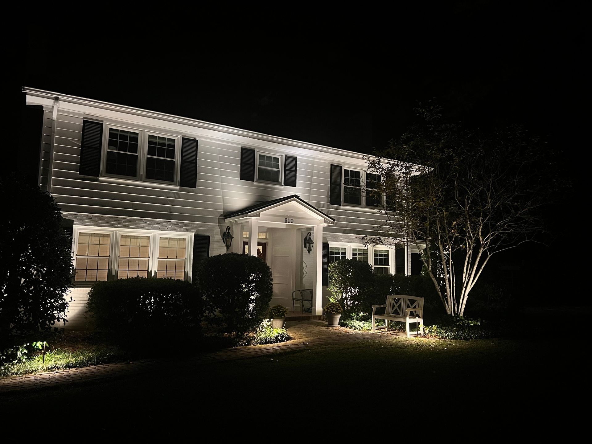 House at night with bright exterior lights and a lit front porch, surrounded by bushes and a bare tree