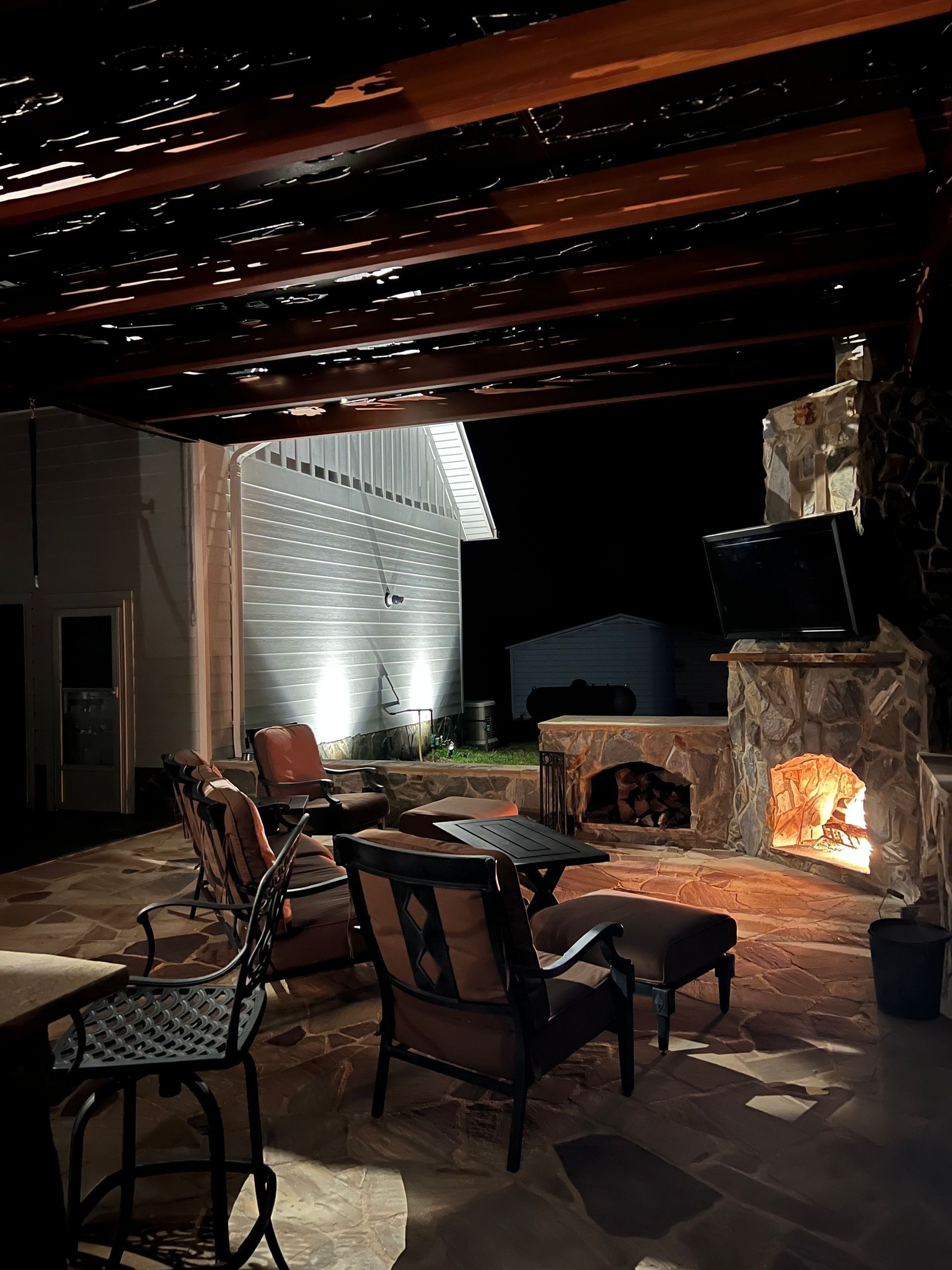 Covered patio with wicker chairs, stone fireplace, and a glowing fire at night