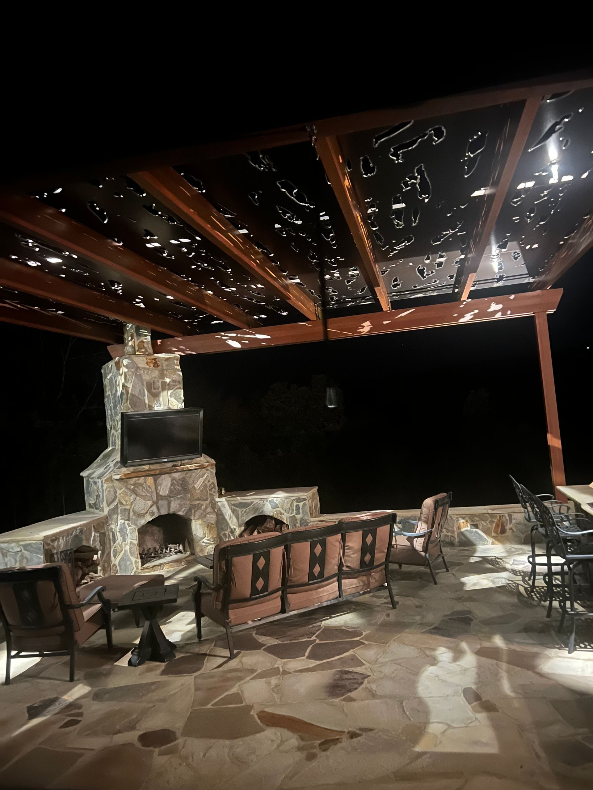 Covered stone patio with outdoor fireplace, wooden pergola, string lights, and seating at night