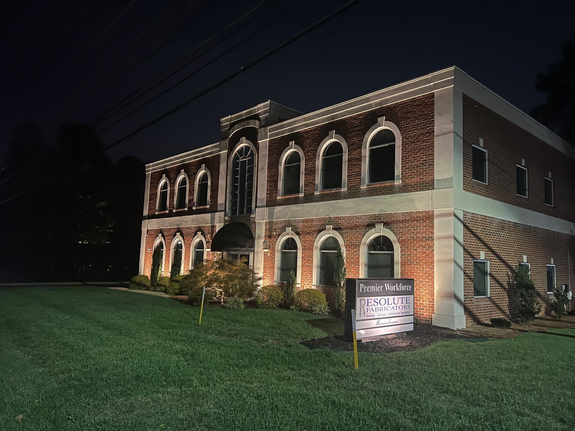 Two-story brick building lit at night, with arched windows and a sign in front on a grassy lawn.