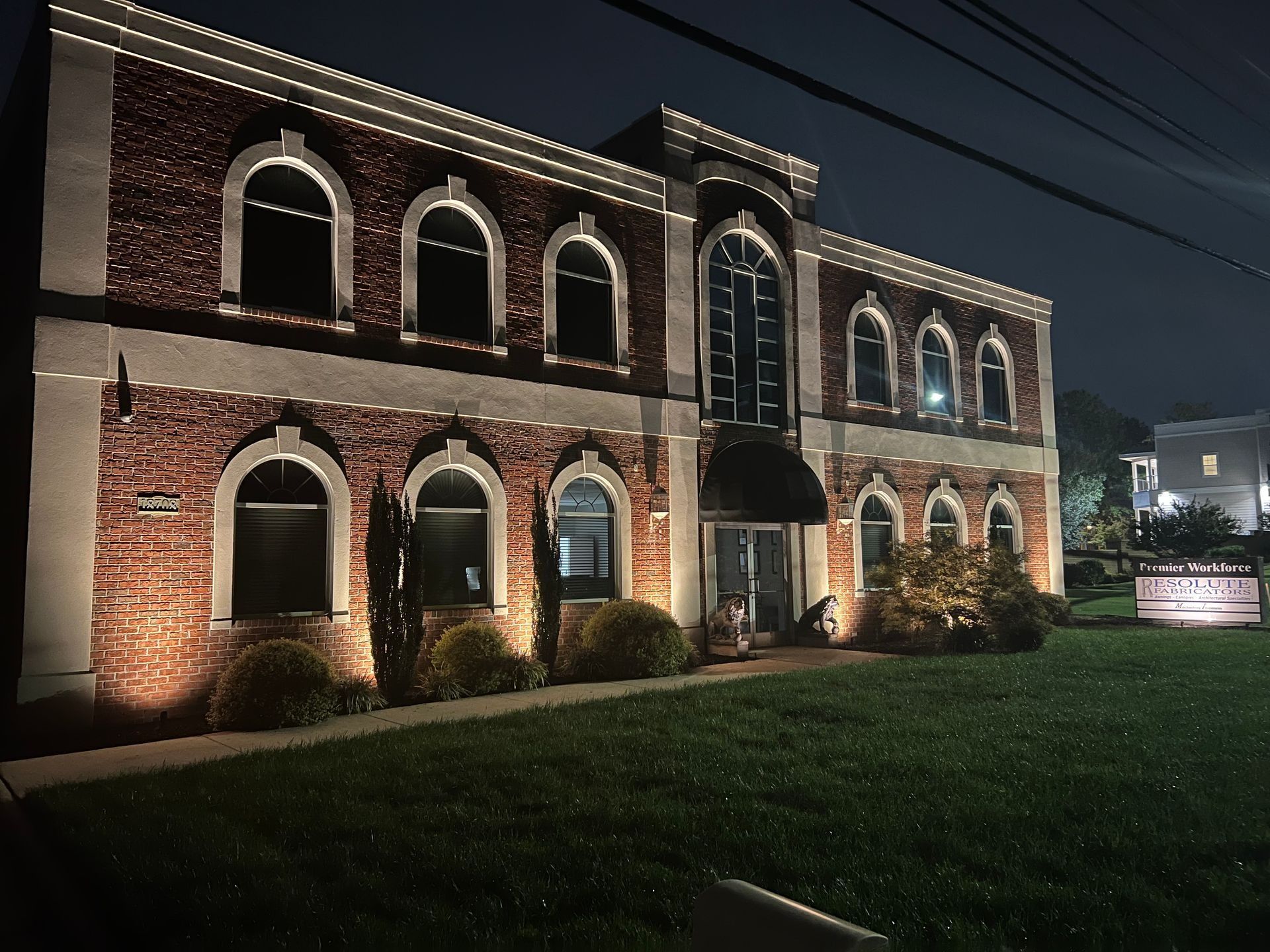 Two-story brick building with arched windows lit at night beside a grassy lawn