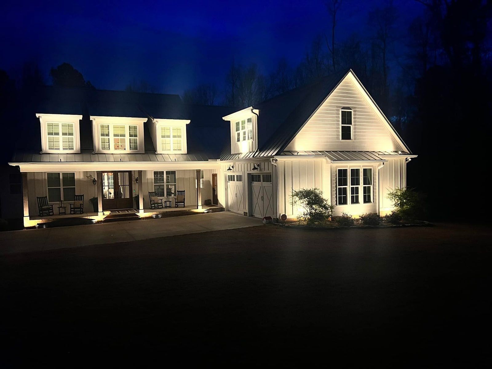 Well-lit white house at night with a wraparound porch and dormer windows