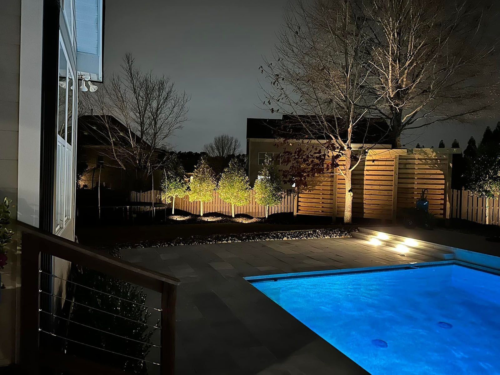 Nighttime backyard with a lit blue swimming pool, wooden fence, and trees beside a house