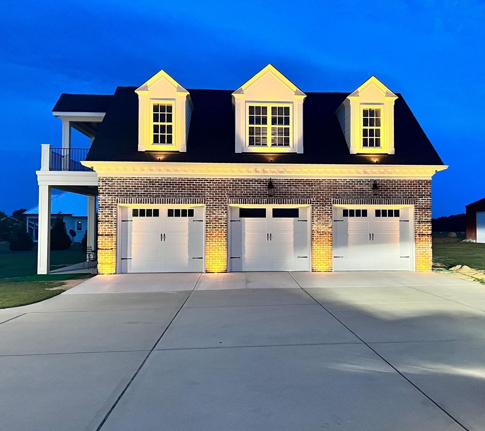 Brick house with three garage doors and dormer windows, lit warmly at dusk under a blue sky