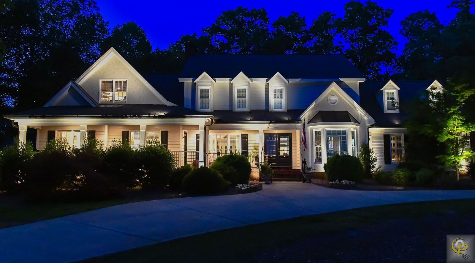 Large illuminated suburban house at night with blue sky, white trim, and trees surrounding the driveway.