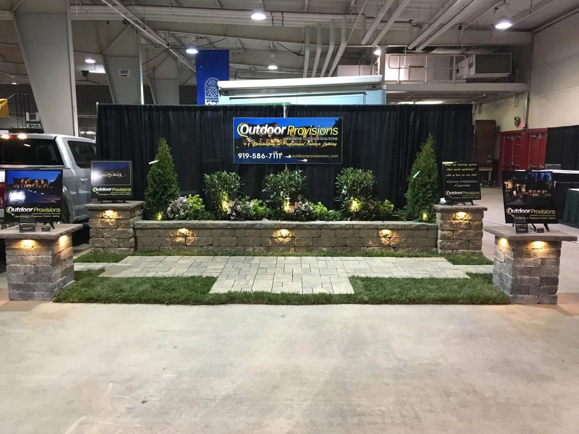 Trade show booth with stone wall, black backdrop, and illuminated sign above plants and turf.
