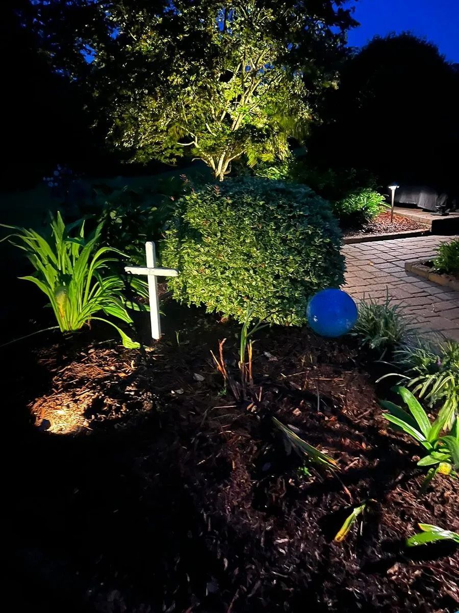 Night garden with illuminated shrubs, a white cross, and a lit brick path.