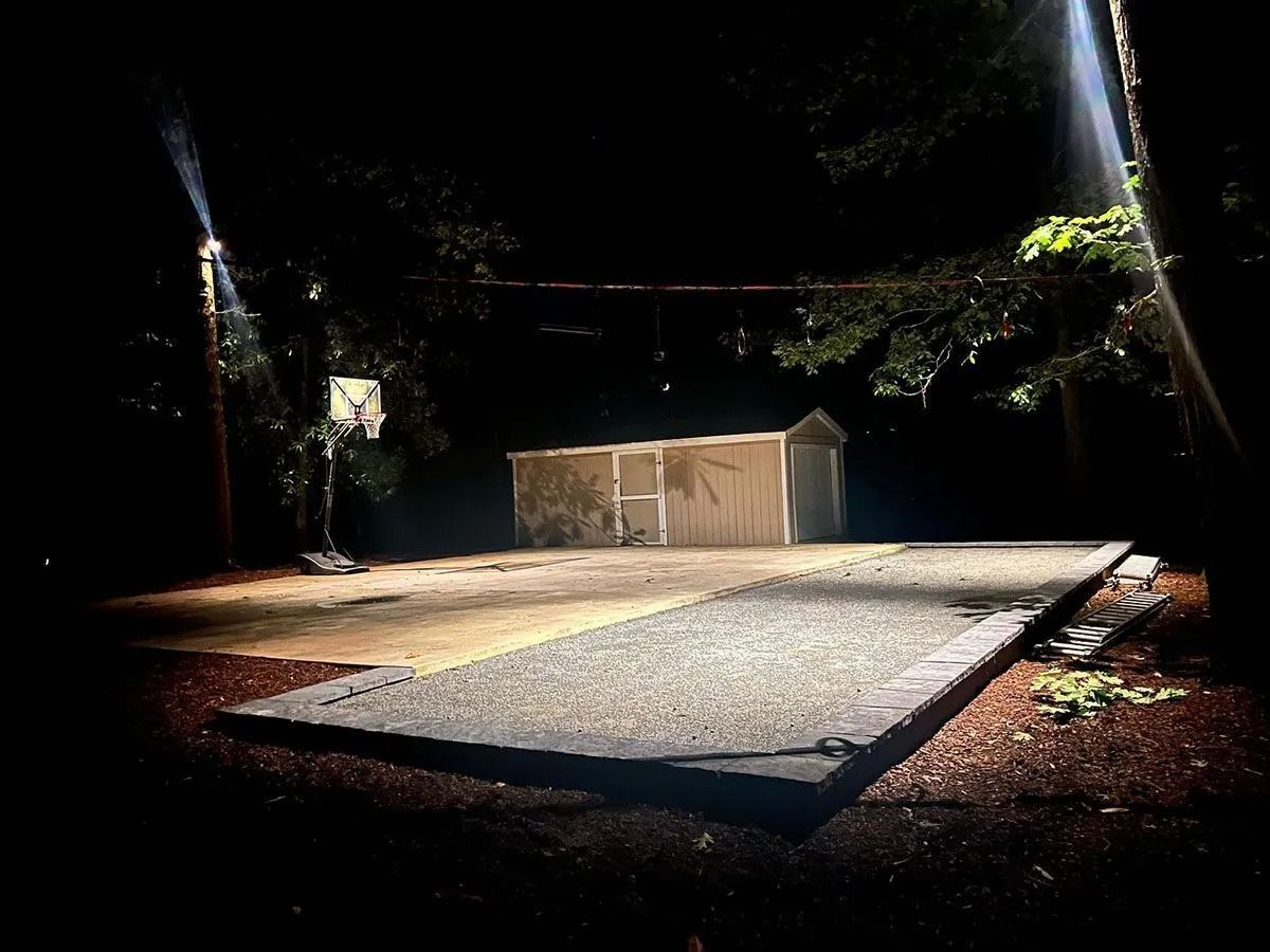 Nighttime outdoor scene with a lit concrete court, small shed, and trees in the background