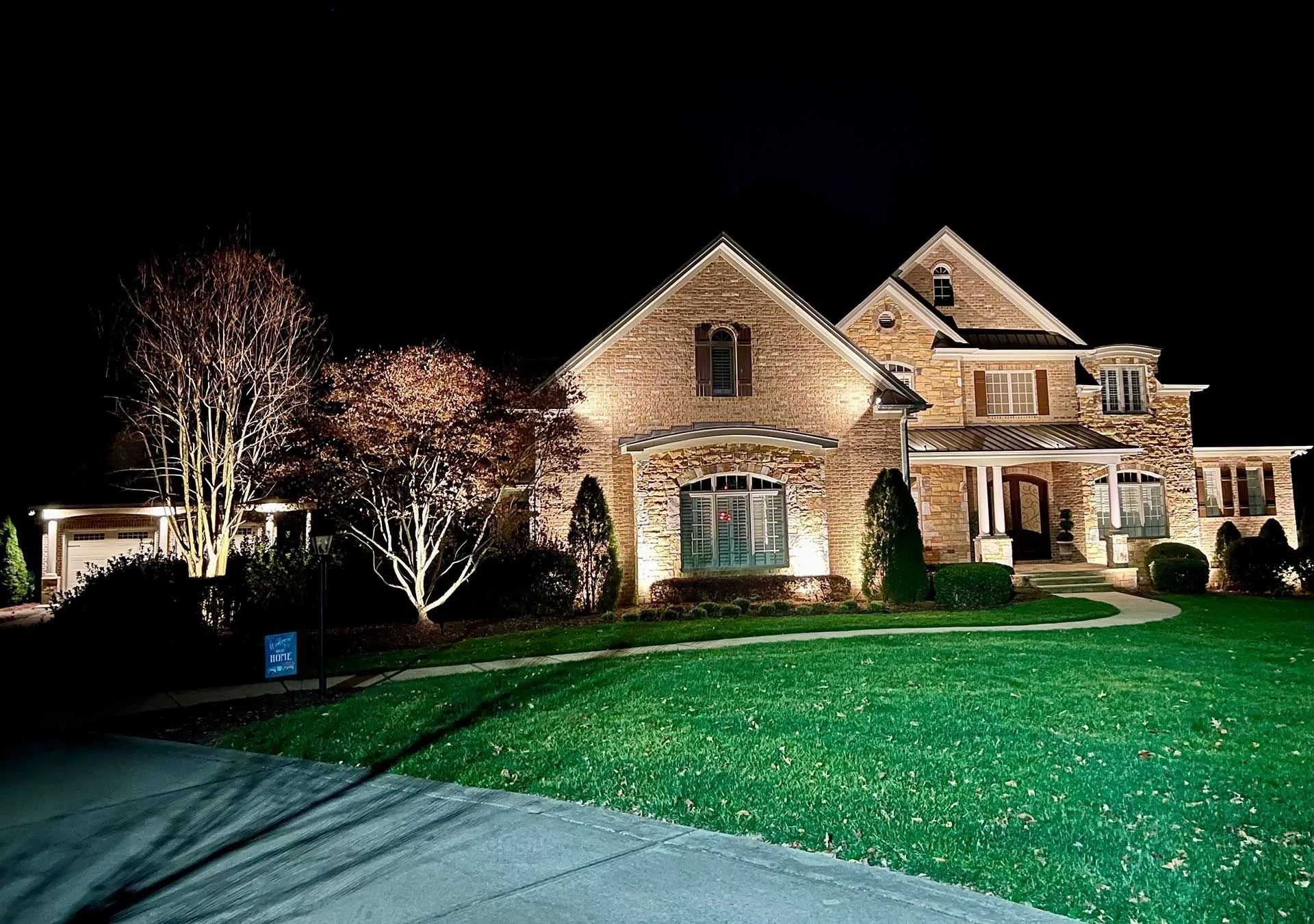 Well-lit stone house with lit trees and a green lawn at night