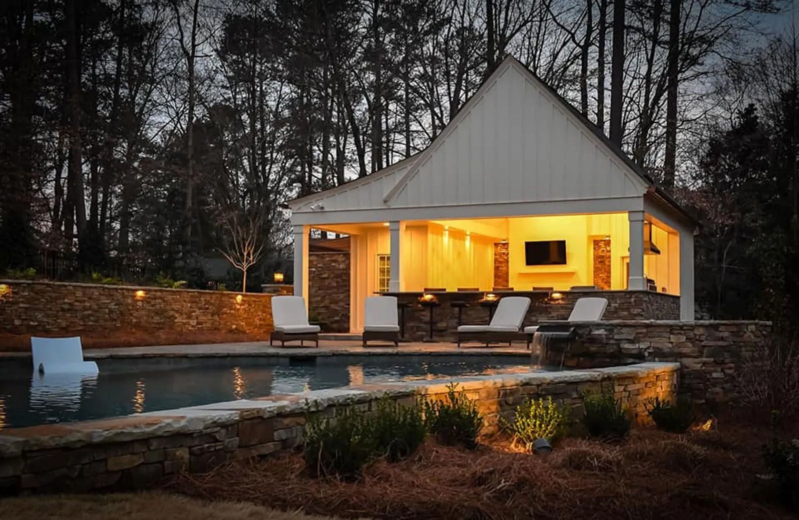 Modern house with a lit covered patio beside a pool at dusk, surrounded by trees and stone landscaping