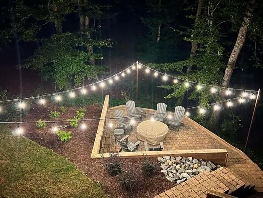 String lights over a backyard patio with fire pit, table, and chairs at night