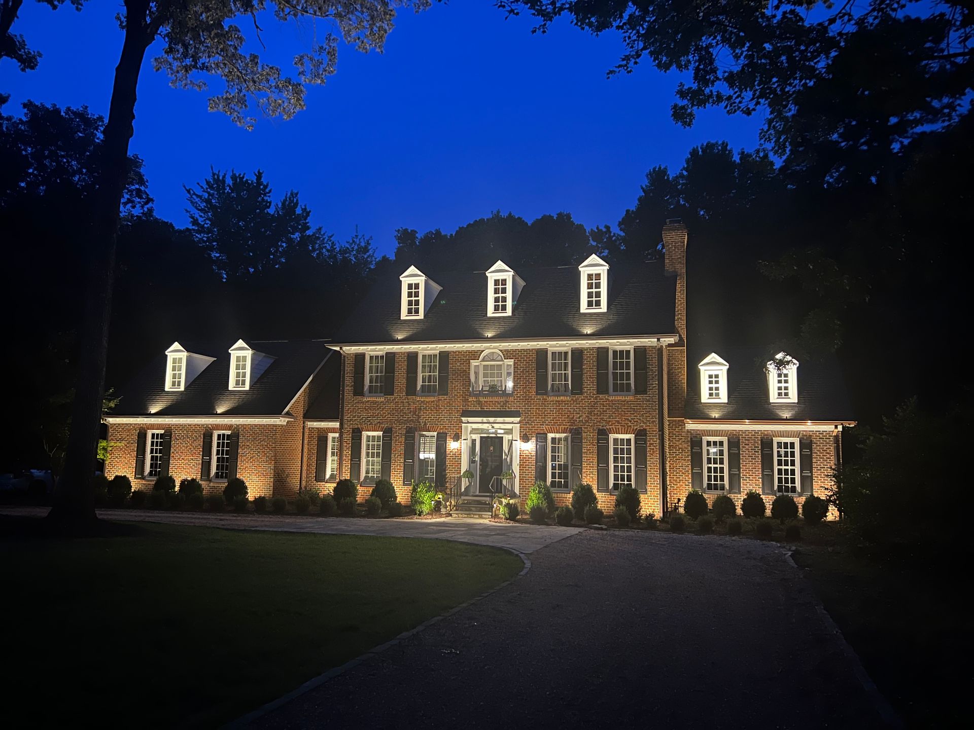 Large brick mansion lit at night, with dormer windows and a dark driveway under a deep blue sky.